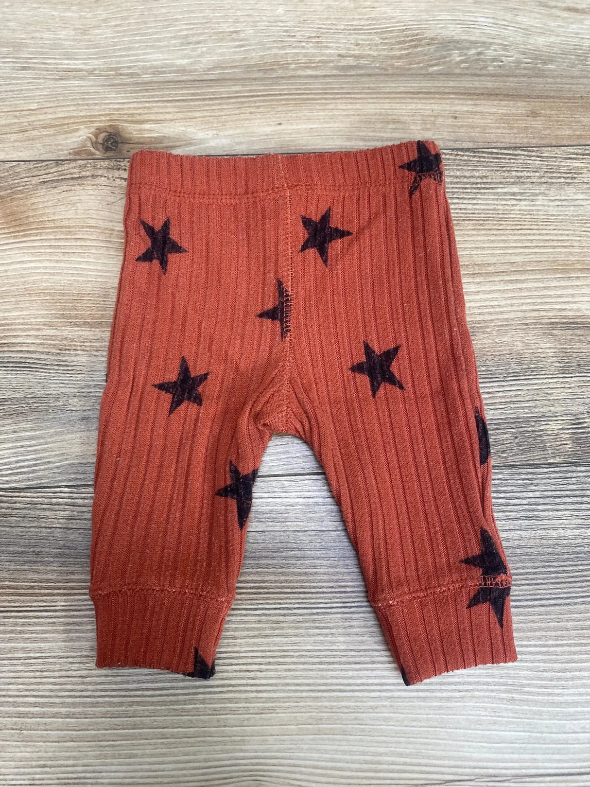 Front view of Grayson Collective ribbed newborn pants in rust showing black stars, elastic waist and cuffs on wood surface