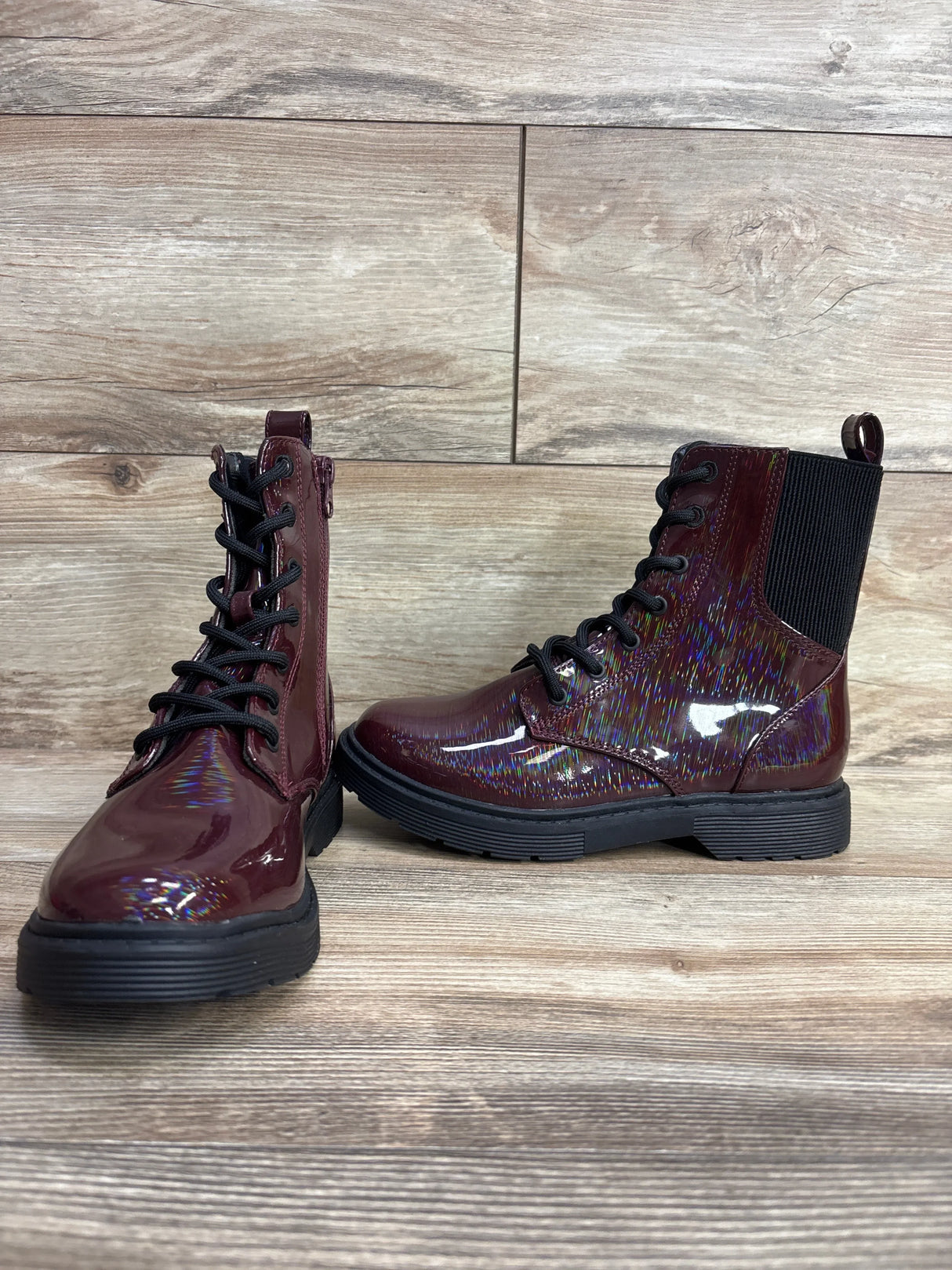 Burgundy kids' lace-up combat boots with iridescent shimmer and black soles on wood floor