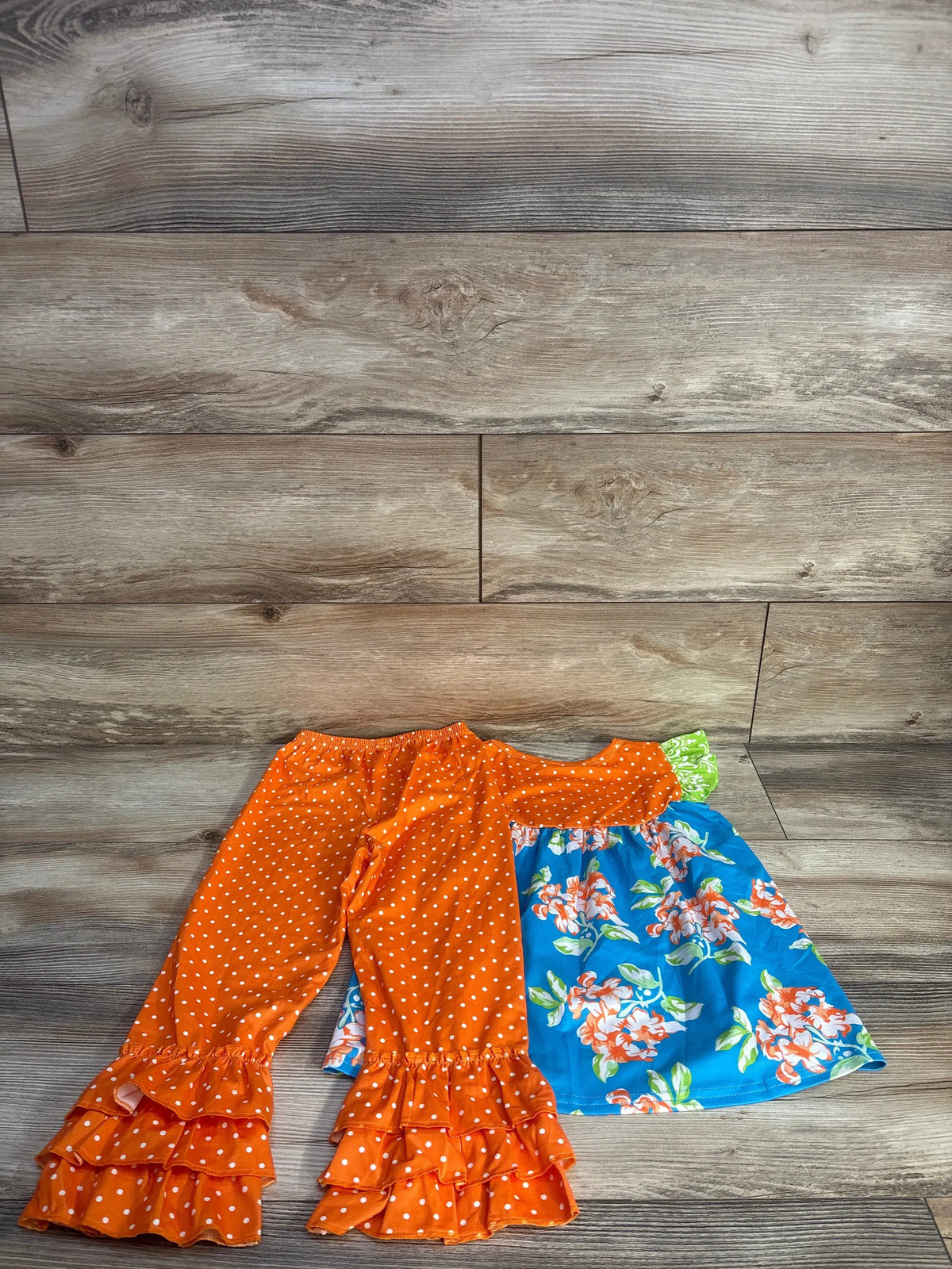 Flat lay of two-piece dress and ruffle pants set showcasing orange polka dot ruffles and blue floral fabric on wood flooring