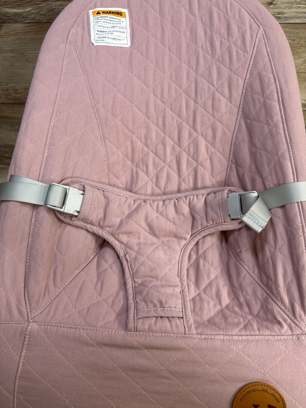 Close-up front of ANGELBLISS baby bouncer in pink showing quilted seat and center harness with buckle on wood floor