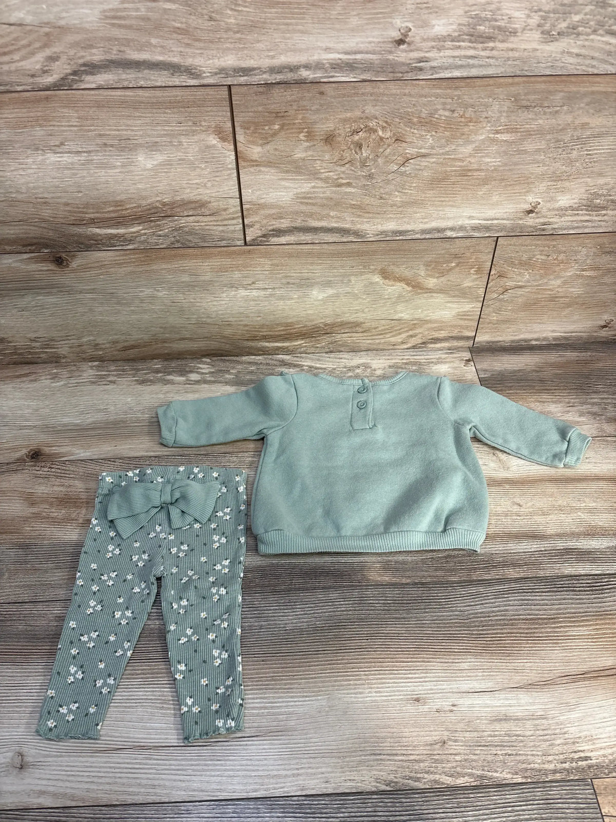 Flat lay of Nicole Miller 2-piece sweatshirt set showcasing sage green sweatshirt with back-button detail and matching floral leggings with bow on wood floor