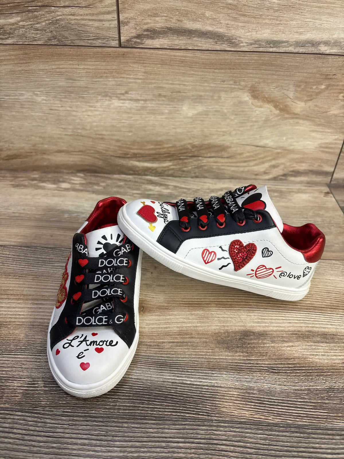 Top view of white Dolce and Gabbana Portofino DG Amore calfskin sneakers with black laces and red heart graphics on wood flooring