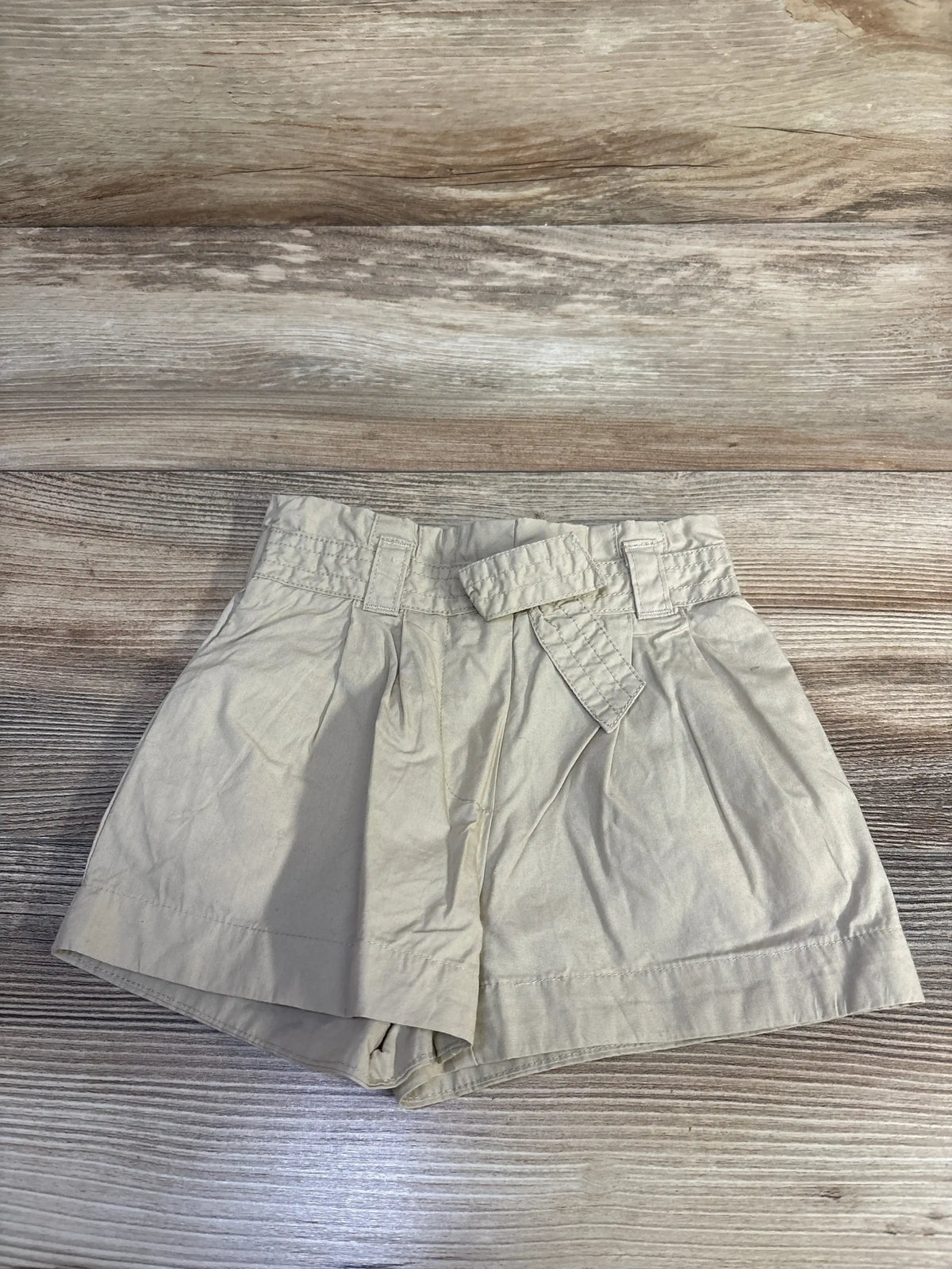 Flat-lay of Janie & Jack beige baby shorts showcasing pleated front, stitched belt detail and cuffed hem on wood background