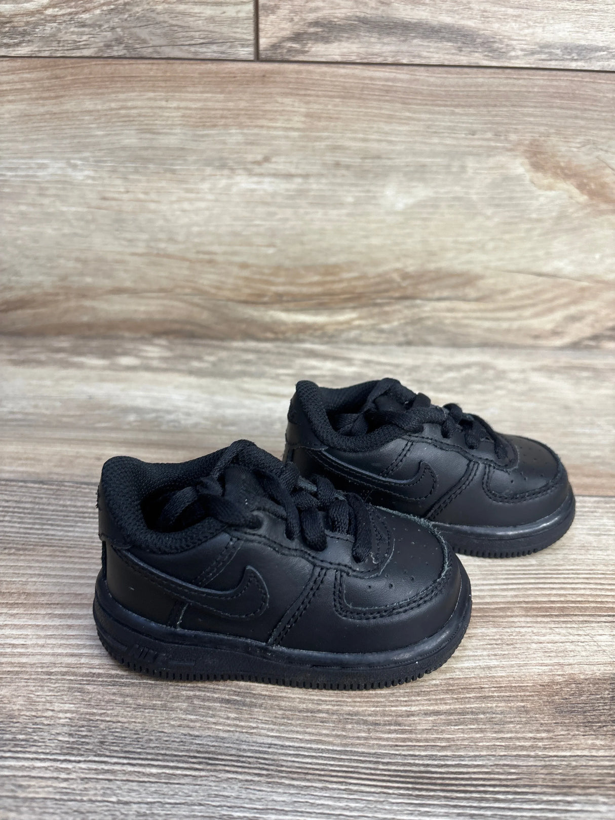 Three-quarter view of Nike Air Force 1 Triple Black toddler sneakers showcasing all-black leather upper and swoosh on wood plank floor