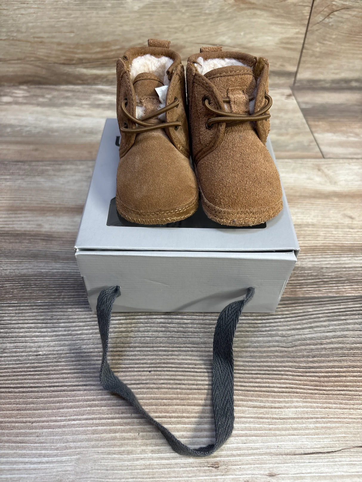 Front view of Ugg Neumel baby booties, chestnut suede with faux shearling lining and laces on a gray box