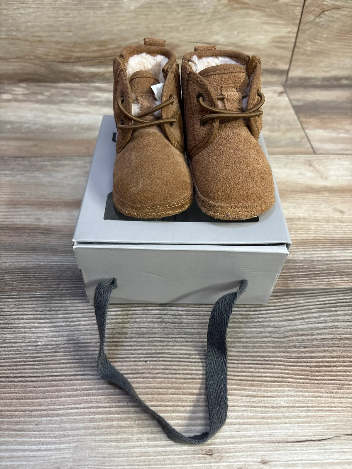 Front view of Ugg Neumel baby booties, chestnut suede with faux shearling lining and laces on a gray box