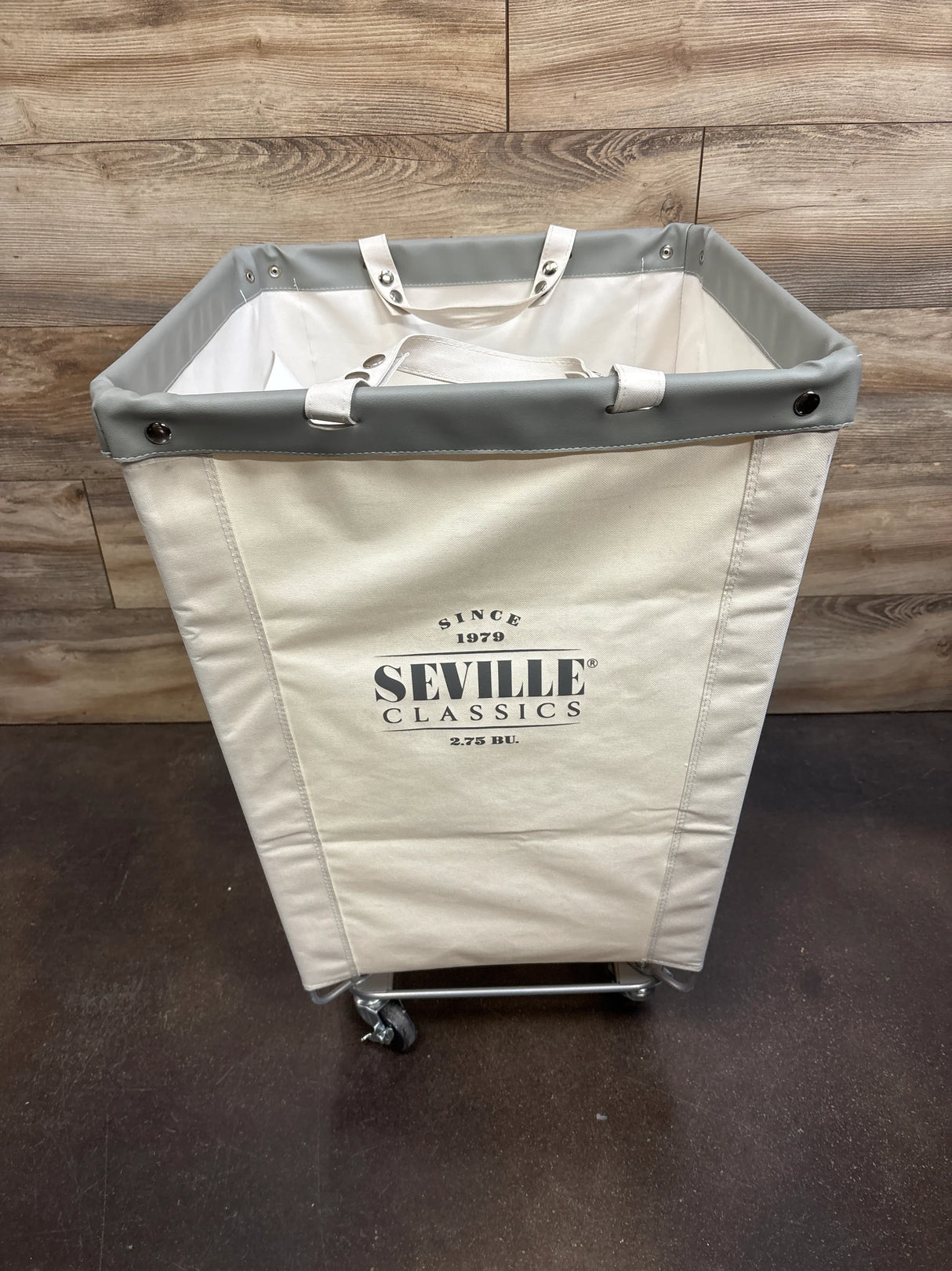 Front view of Seville Classics rolling canvas hamper showing gray trim, printed logo, and caster wheels on wood floor