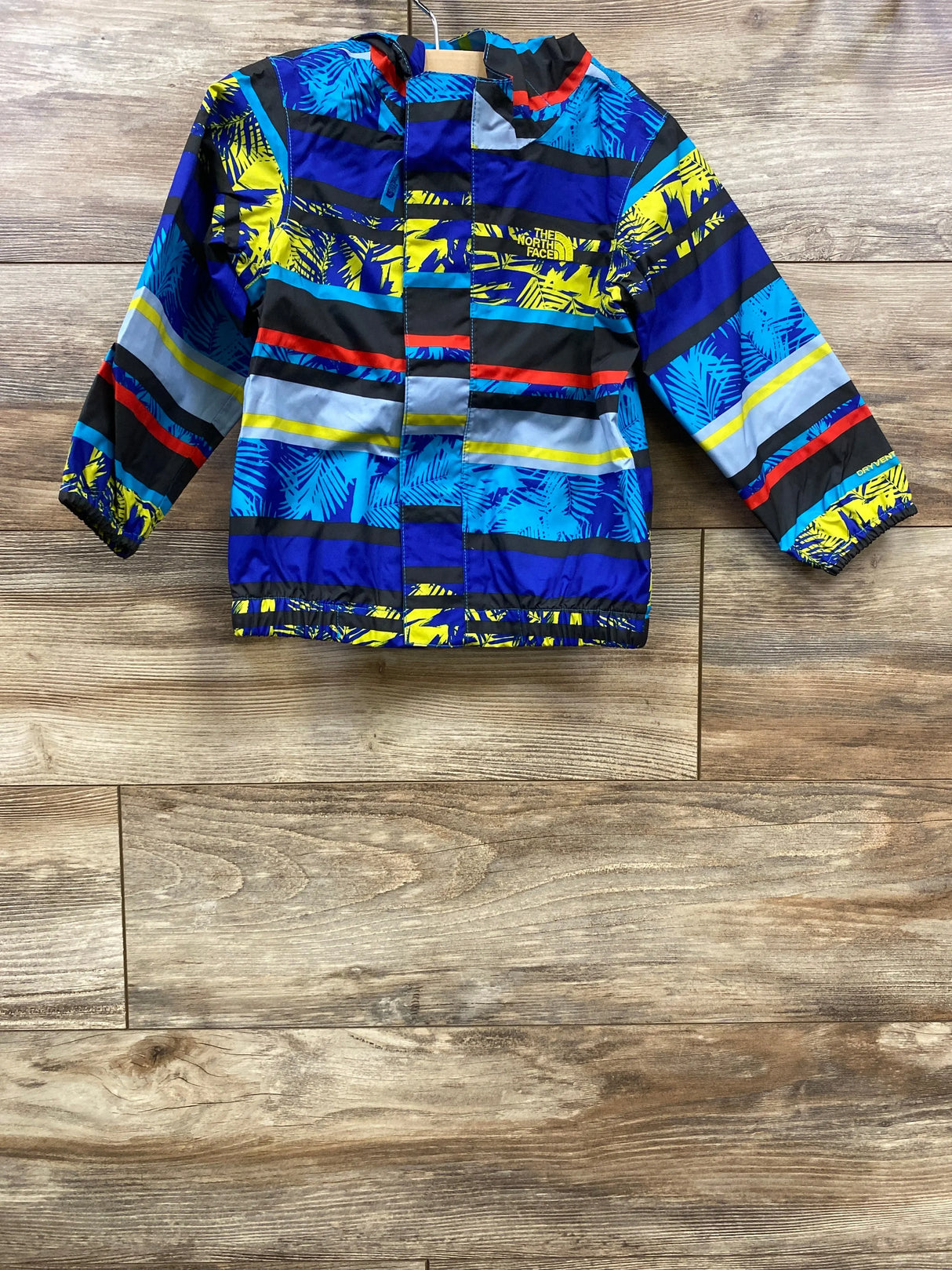 Front view of The North Face Tailout toddler rain jacket showcasing blue, navy, yellow and red palm-and-stripe print with wood plank floor background