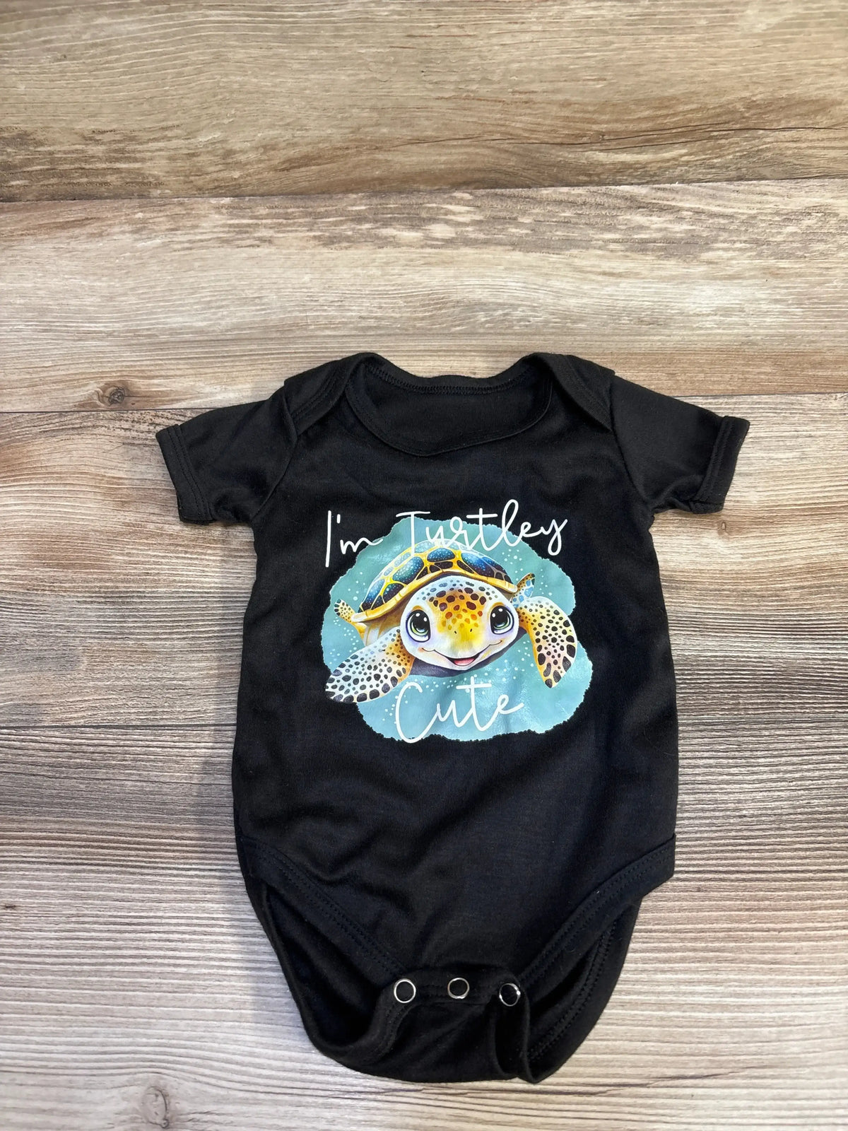Front view of black 3-6m baby bodysuit with sea turtle graphic and text I’m Turtley Cute on wood background