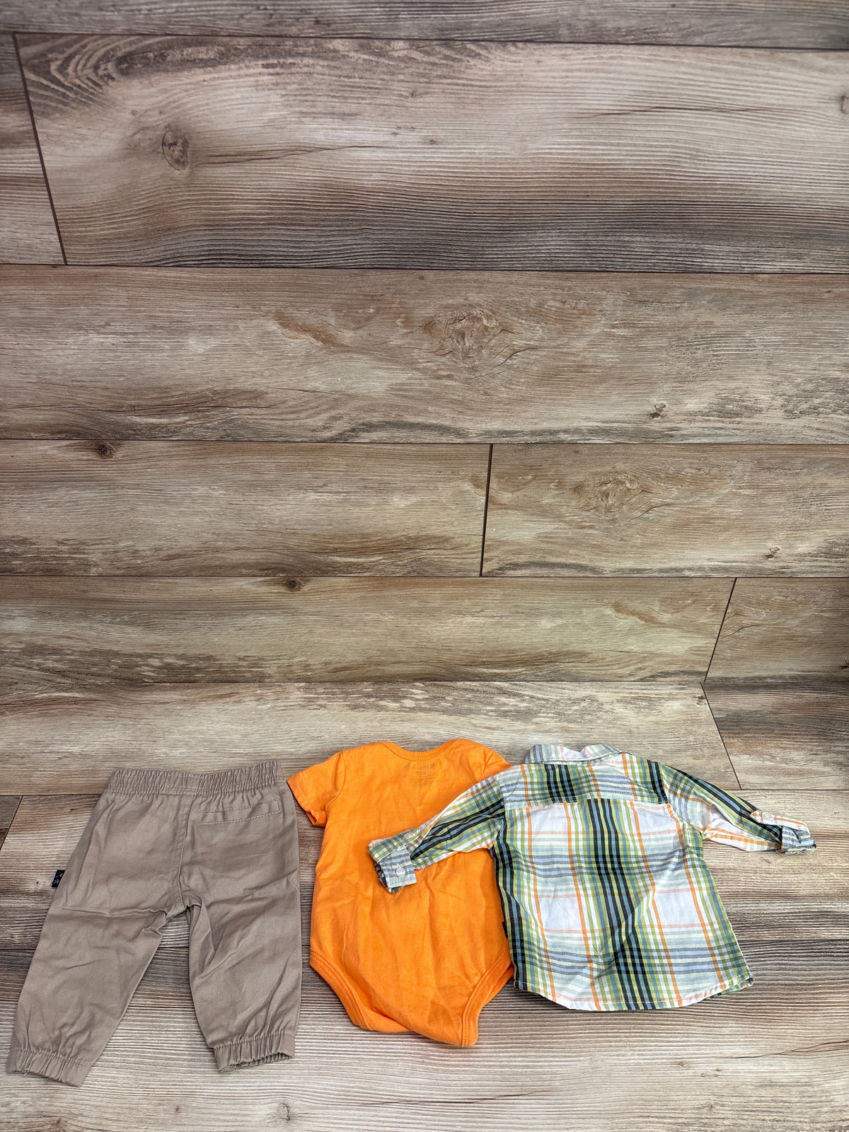 Top view of American Hawk three-piece baby set with plaid button-up shirt, orange bodysuit, and taupe pants on wood flooring