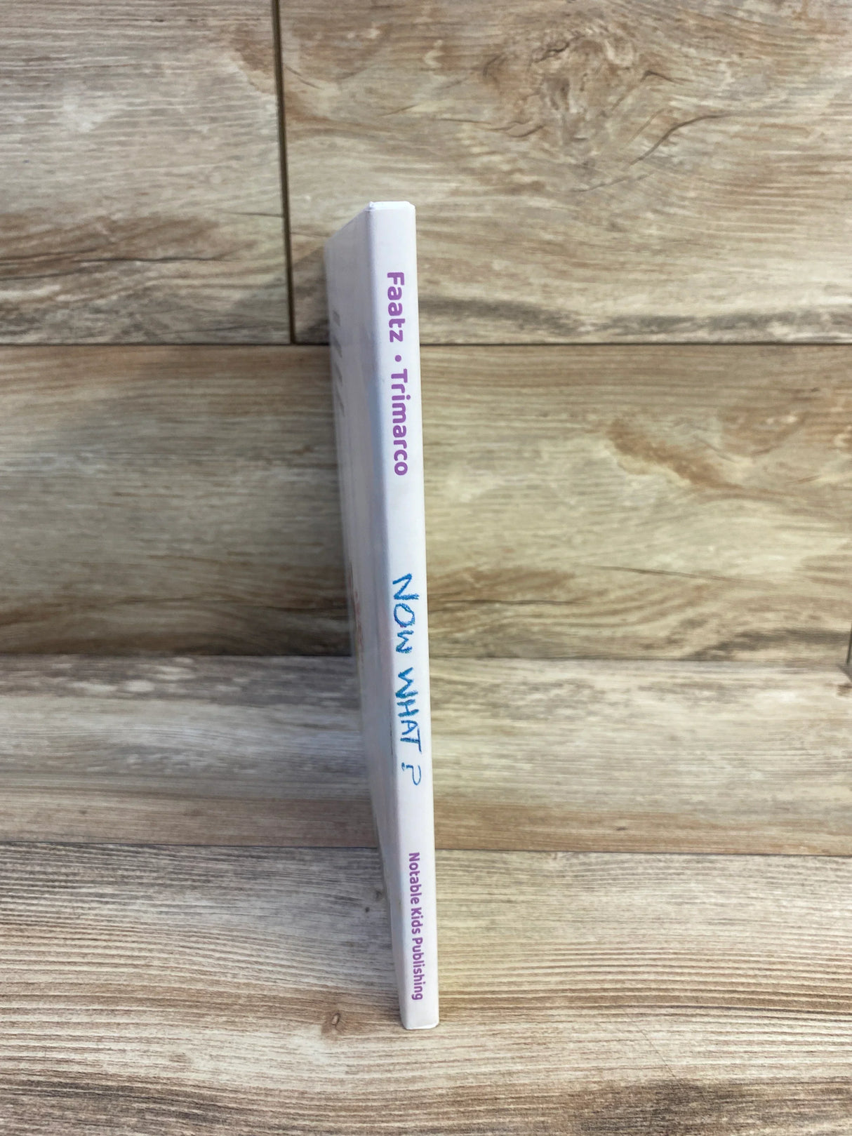 Spine view of Now What hardcover book by Faatz and Trimarco showcasing white spine with Notable Kids Publishing label on wood plank floor
