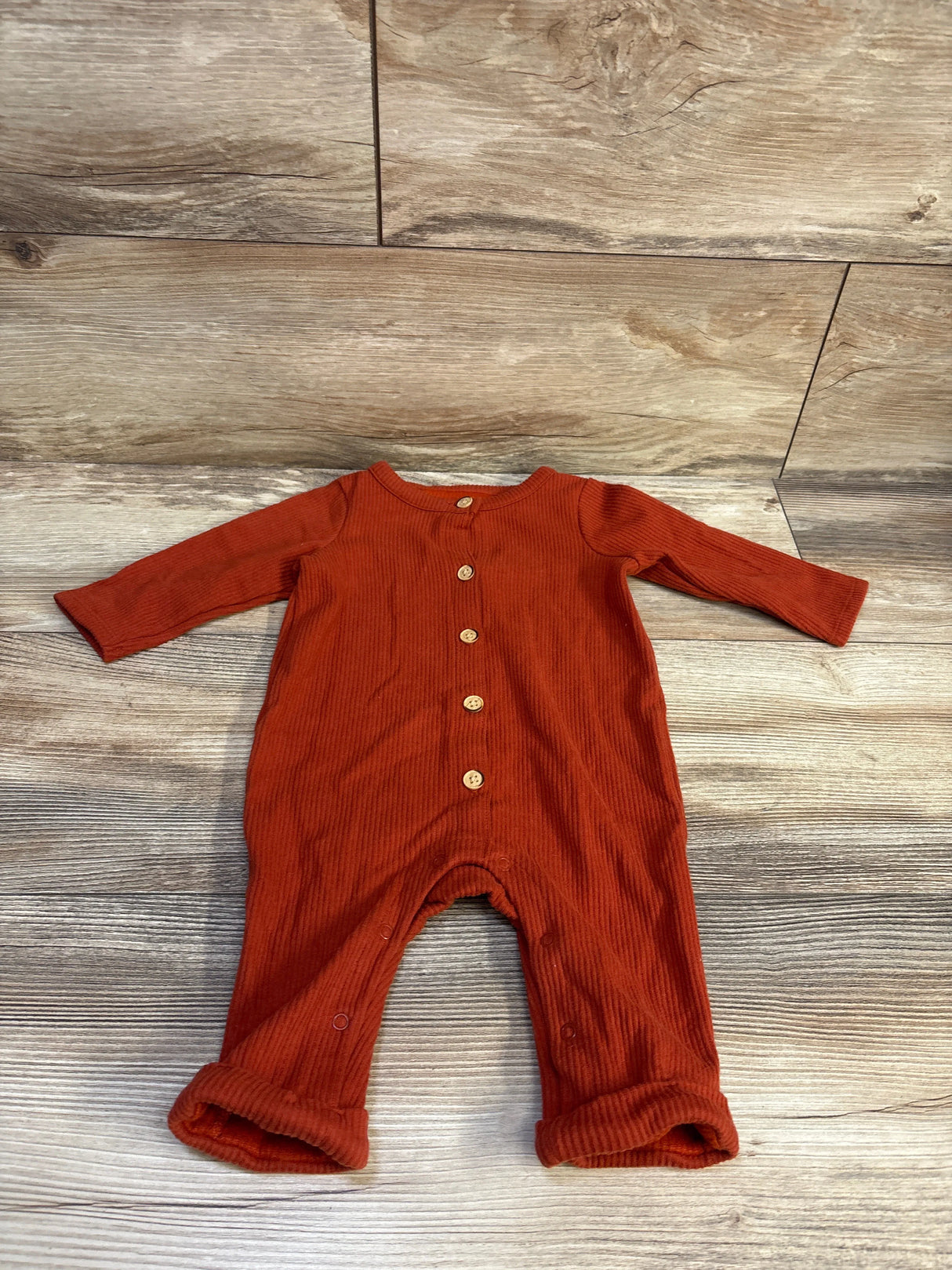 Flat-lay of rust ribbed button-front baby coverall with wooden buttons and leg snaps on light wood background