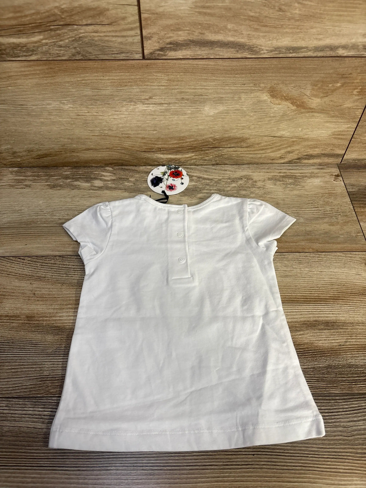 Back view of white Tessa James Birdie baby tee showcasing puff sleeves and snap closures on a wood floor