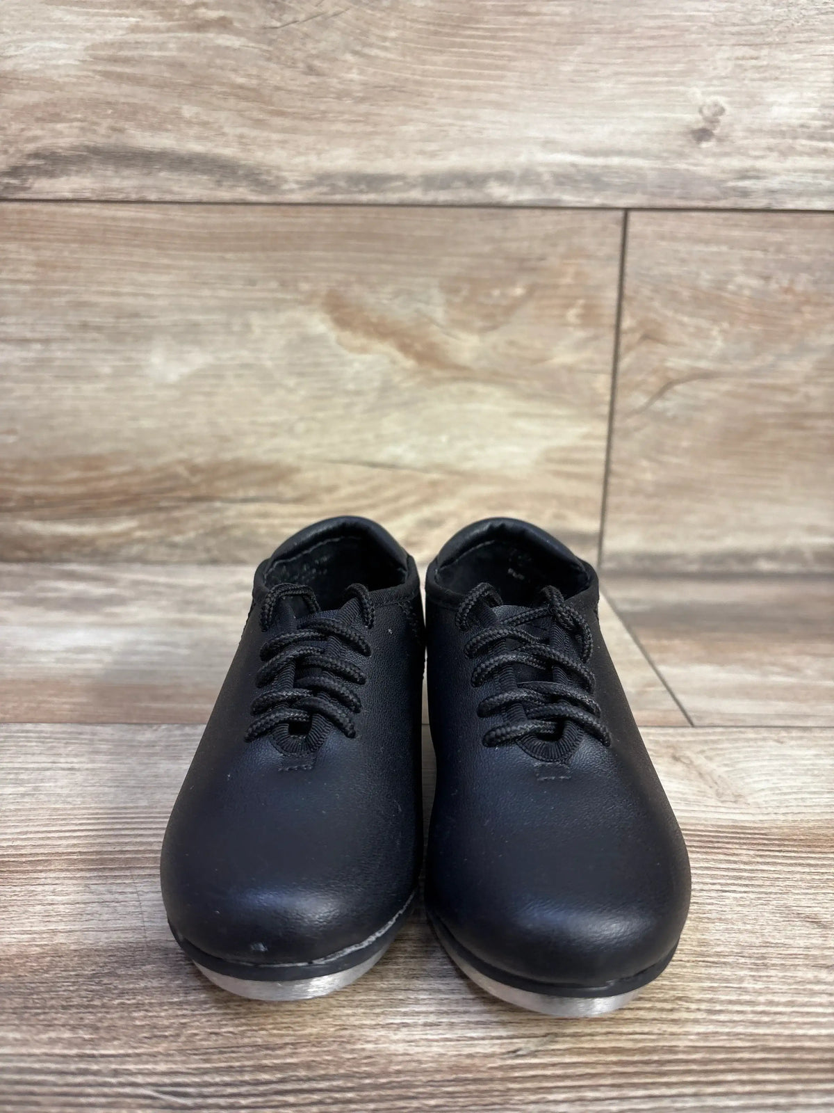 Front view of black Theatricals toddler tap shoes size 7C showing laced front and metal taps on a light wood floor background