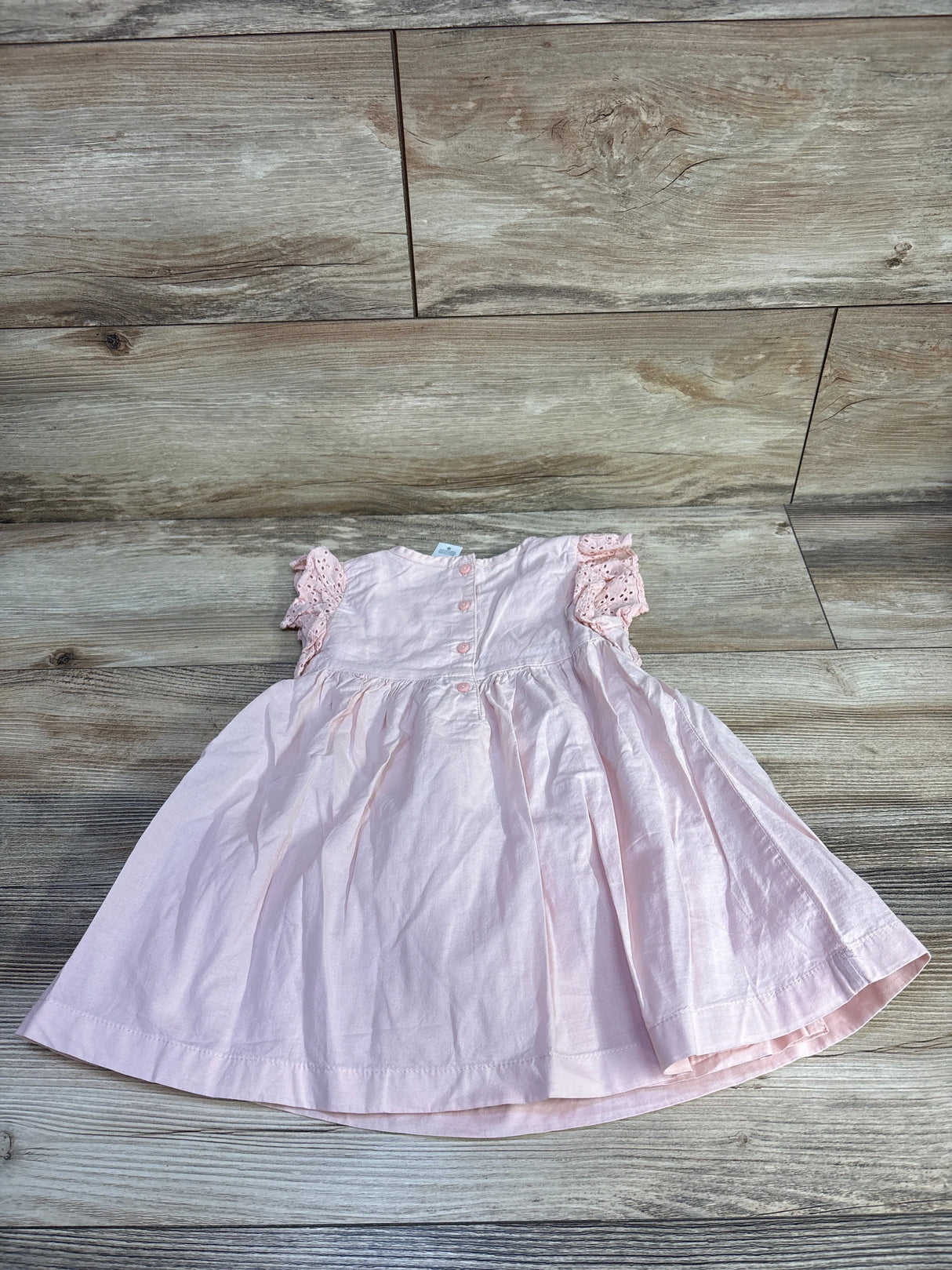 Front-flat view of pink eyelet toddler dress showcasing eyelet flutter sleeves and back button closure on light wood floor background