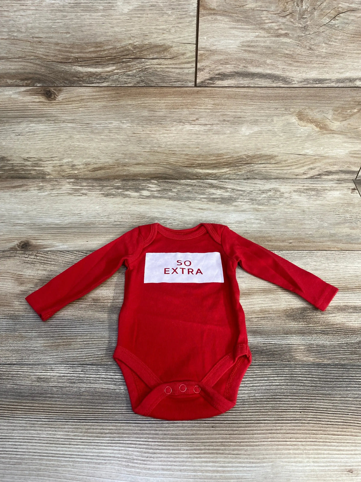 Front flat lay of red newborn long-sleeve bodysuit with white chest panel reading SO EXTRA on wood background