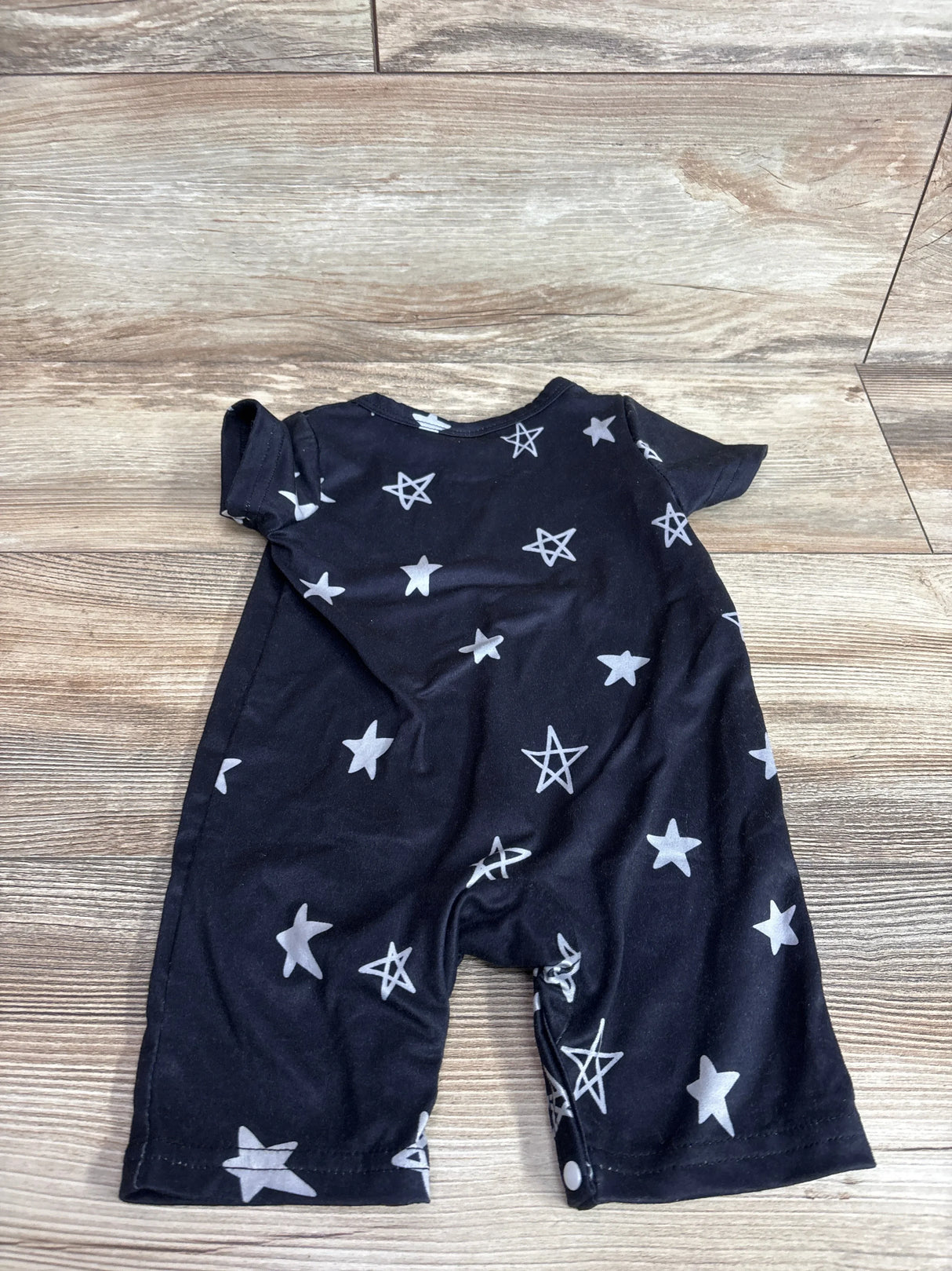 Front flat-lay of black shortie baby romper (6-9 months) showing white star print, short sleeves, and snap crotch on wood floor