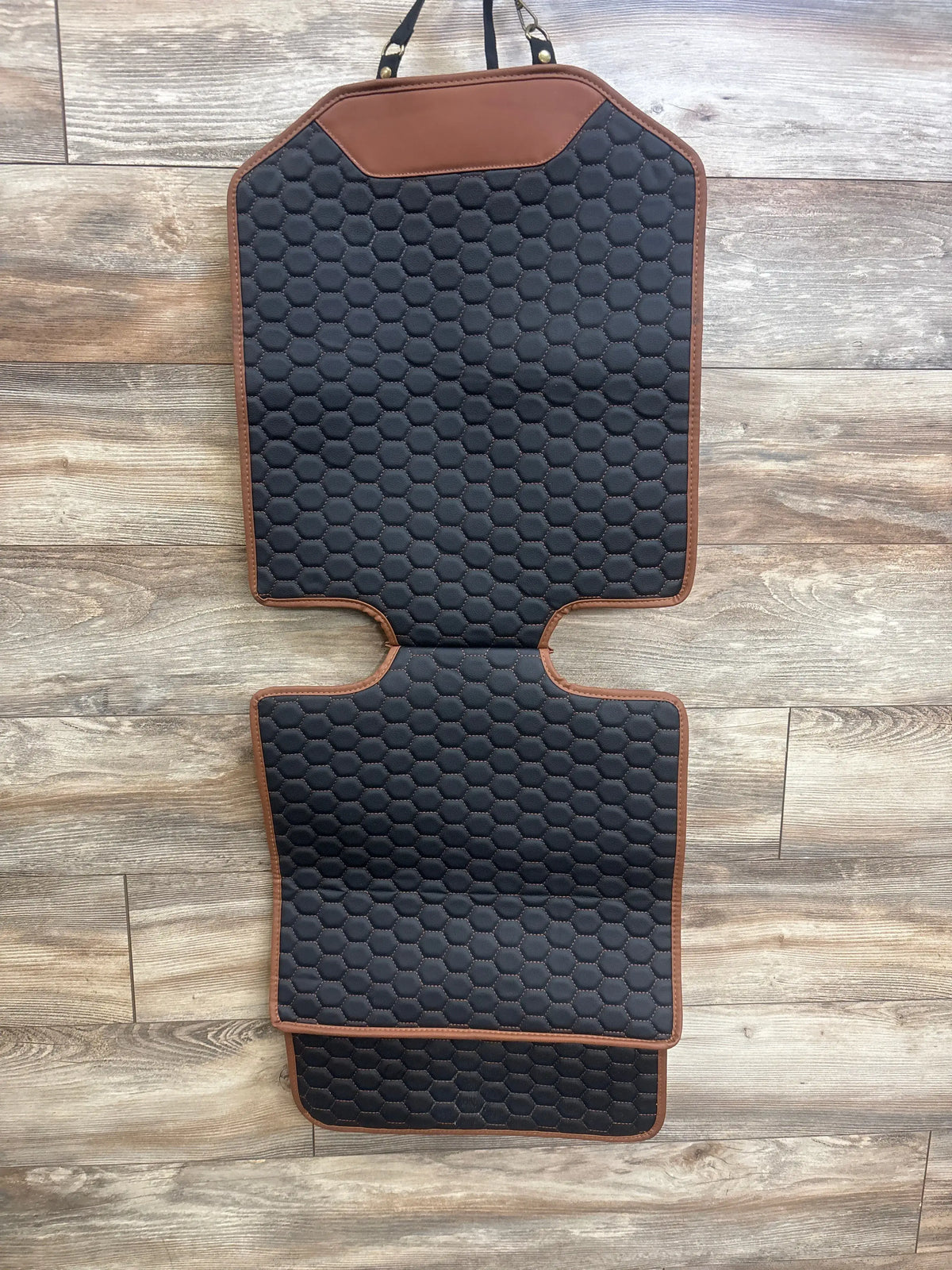 Top view of Owleys Premium Leather Car Seat Protector showcasing black hexagonal quilting, brown trim, and attached straps on wood flooring