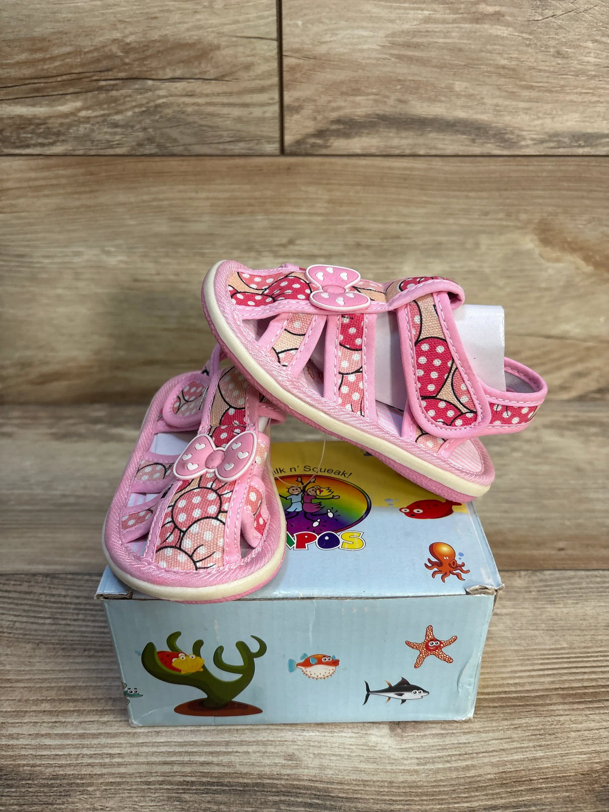 Front-angle of Papos Walk n' Squeak pink toddler sandals, size 6/7C, showing polka-dot bows and Velcro strap on a blue illustrated shoe box