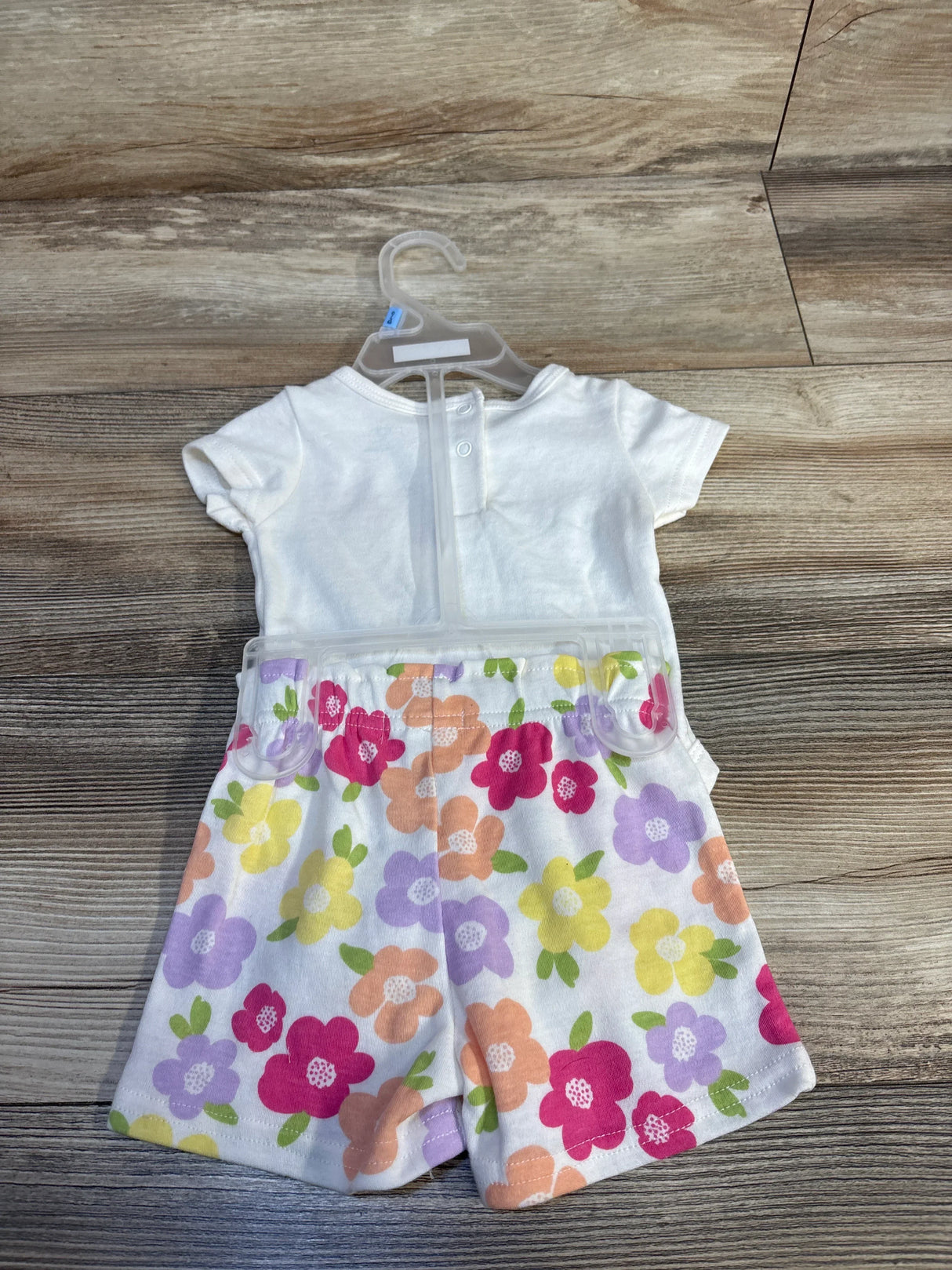 Front view of newborn white short-sleeve bodysuit and floral shorts set showing multicolor flower print and snap closures on wood floor