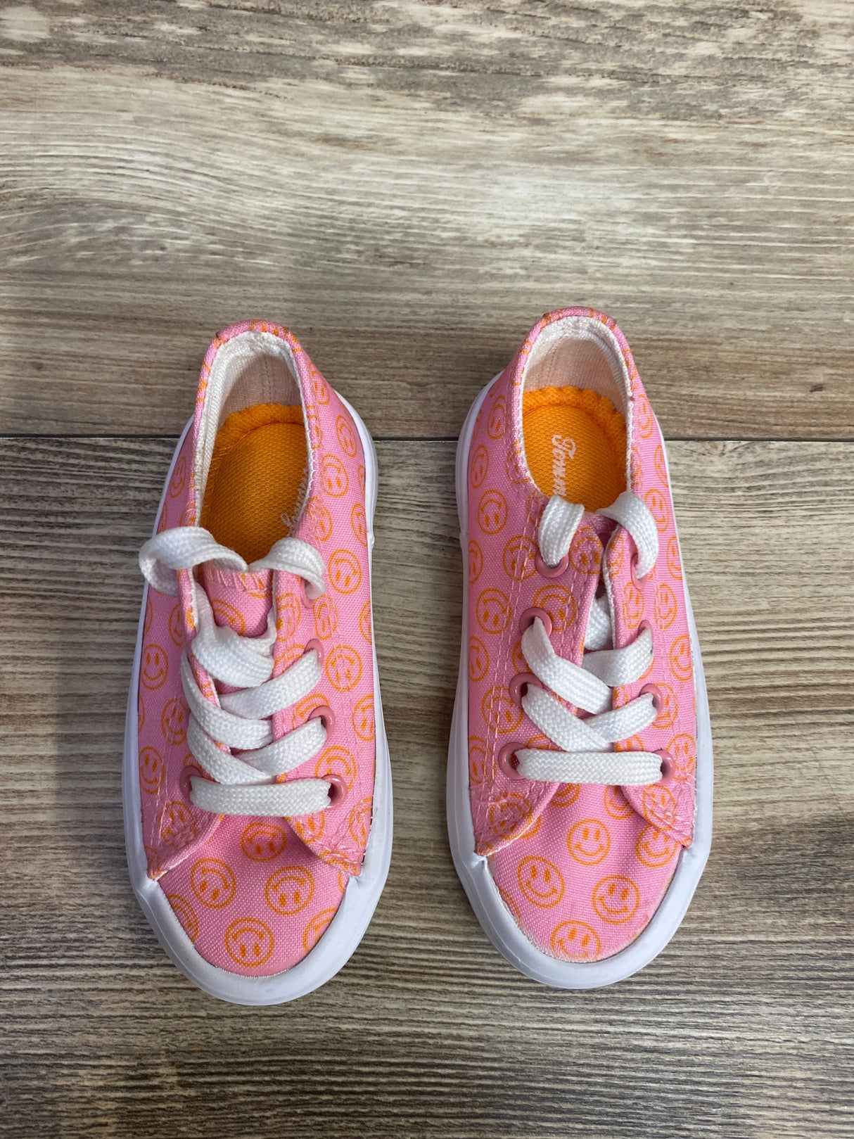 Pair of pink toddler sneakers with orange smiley face print and white laces on wooden floor