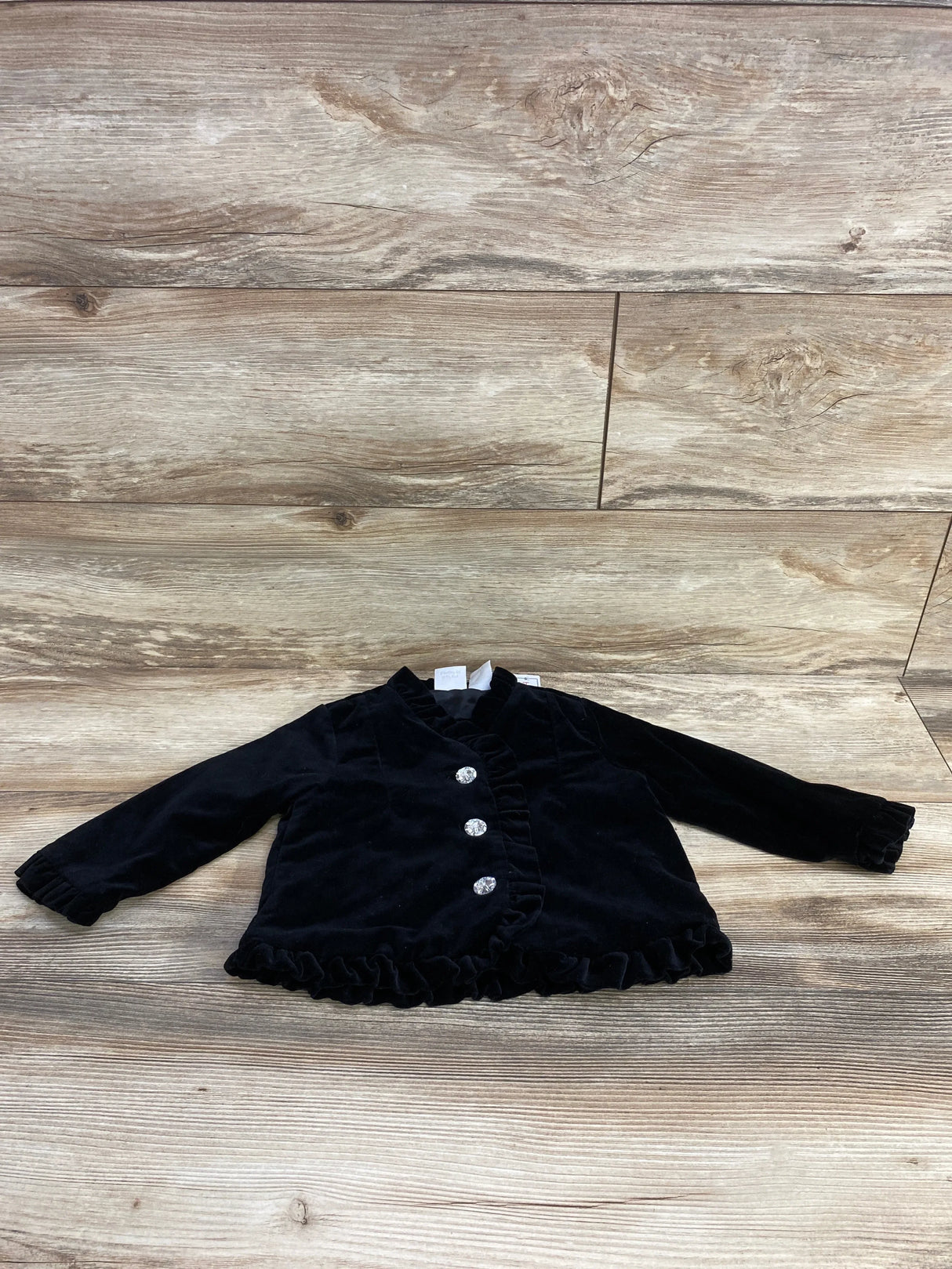 Black velvet baby jacket with ruffled edges and three decorative crystal buttons on wooden background