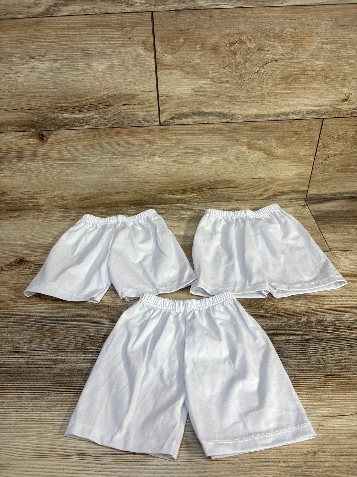 Three pairs of white kids' shorts with elastic waistbands on wood floor, perfect for baby or toddler clothing resale.