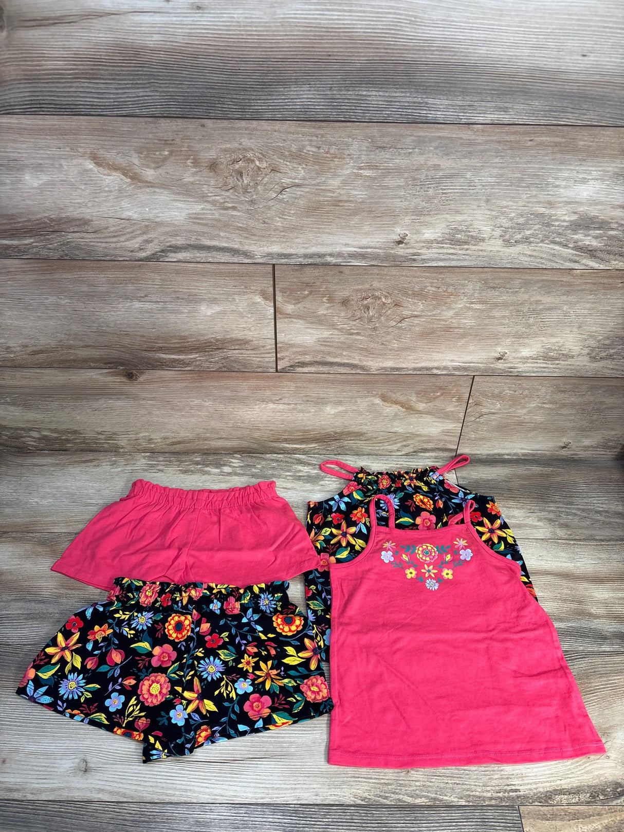 Flat lay of girls' 4-piece set: pink embroidered tank, black floral halter, floral shorts, pink skort on wood floor