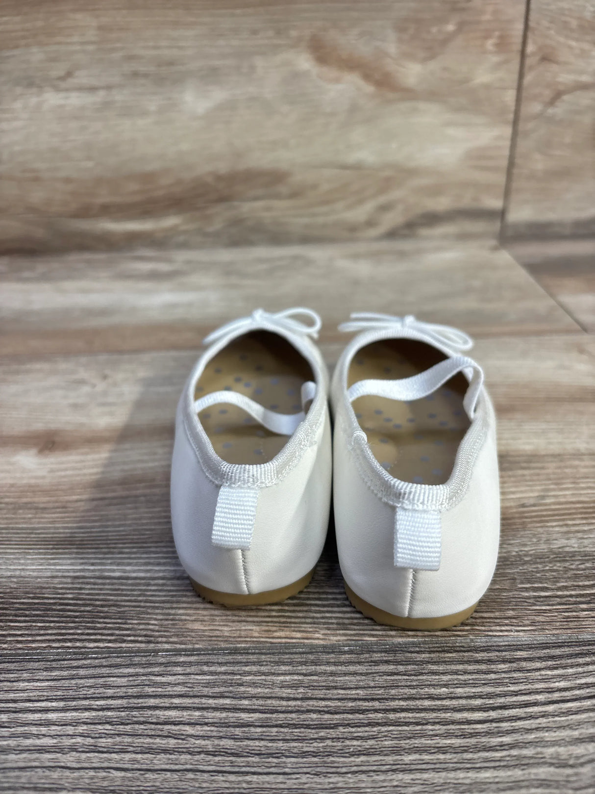 Rear view of Cat & Jack Nora white slip-on ballet flats showing heel pull tab, elastic strap, bow detail and tan sole on light wood background