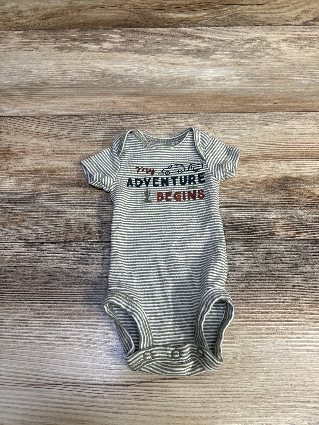 Top-down view of preemie green striped Carter’s bodysuit with ‘My ADVENTURE BEGINS’ text, car and cactus graphics on wood background