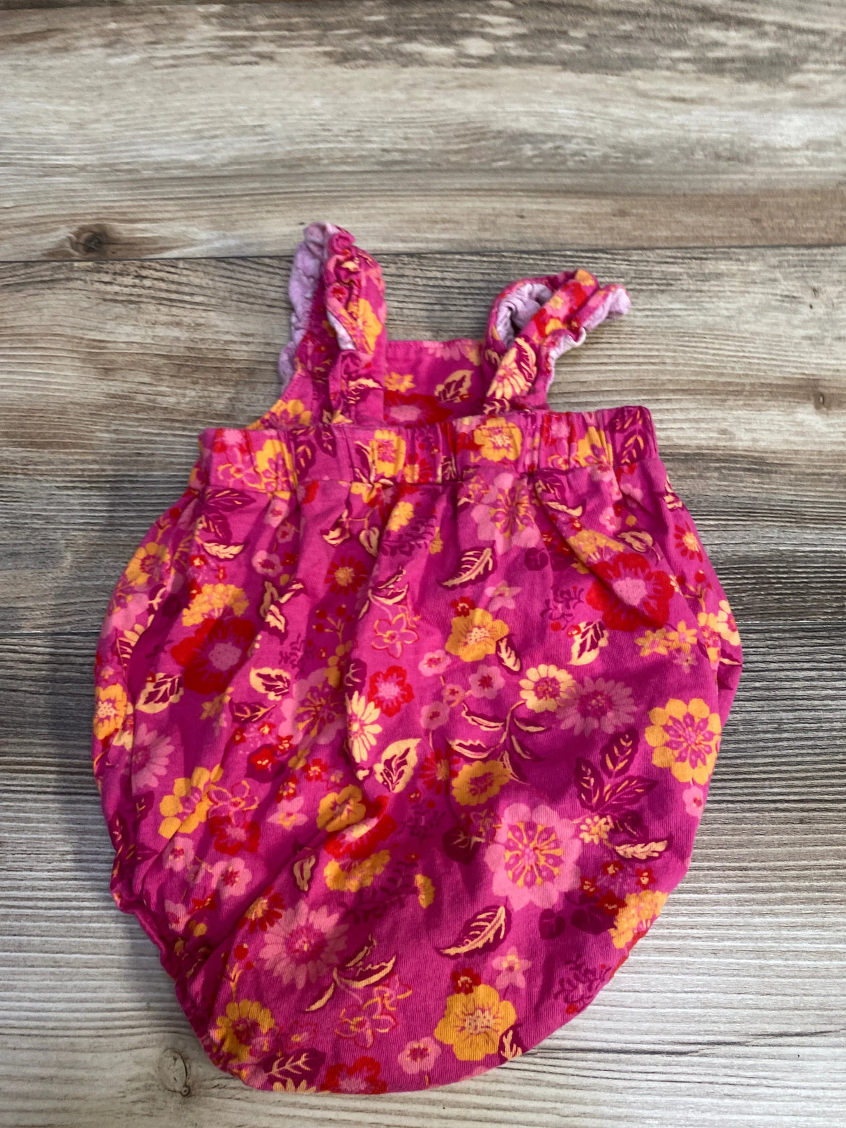 Top-down view of pink floral bubble romper (3-6 months) showing ruffled shoulder straps and elasticized legs on wood