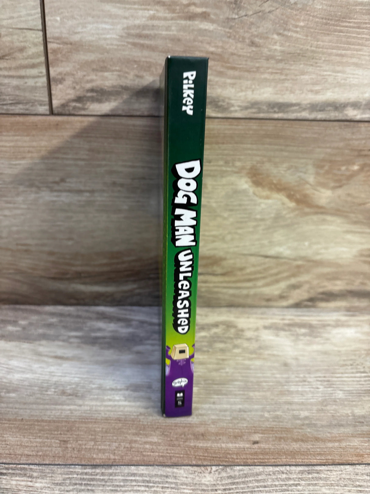 Spine view of Dog Man Unleashed #2 hardcover showing green spine with white title text and purple character on wood background