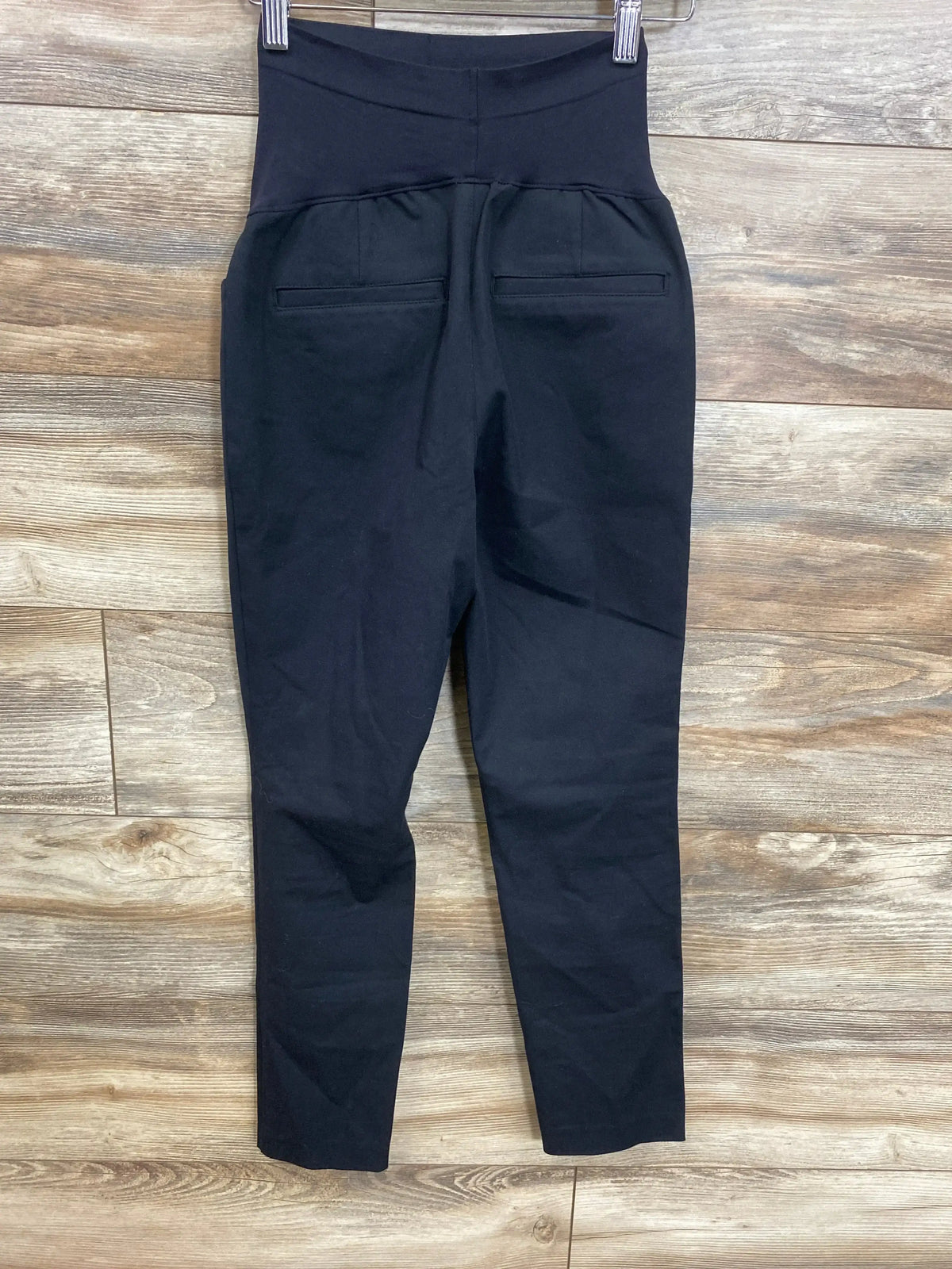Black maternity pants with high waistband and back pockets, hanging on hanger against wood floor