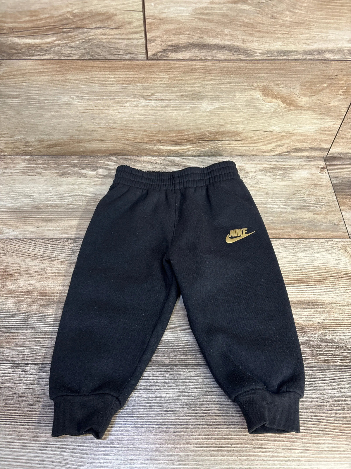 Front view of black Nike joggers showcasing elastic waistband, cuffed ankles, and gold swoosh logo on wood floor