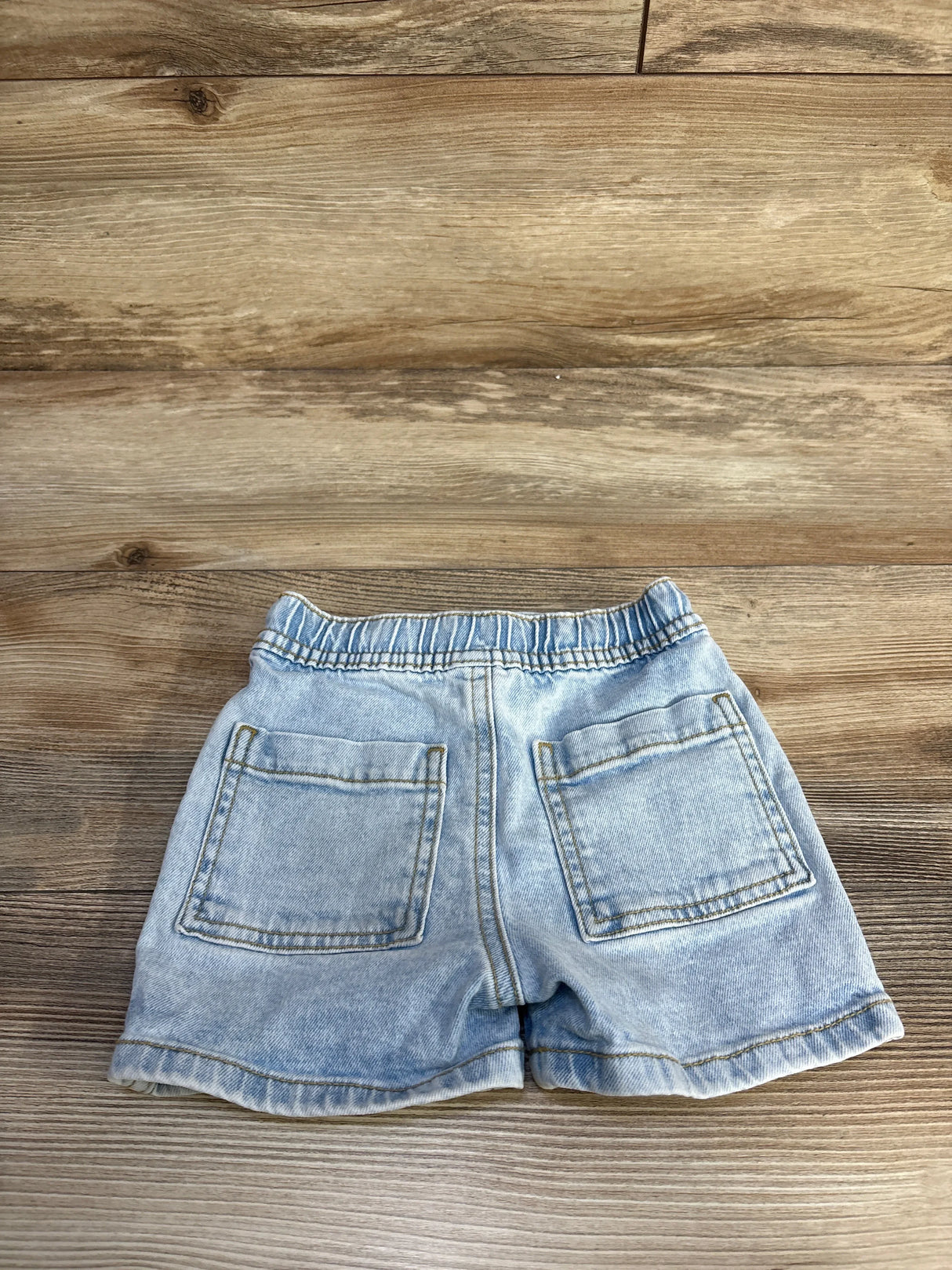 Back view of light blue toddler jean shorts showing elastic waistband and two patch pockets on wood background