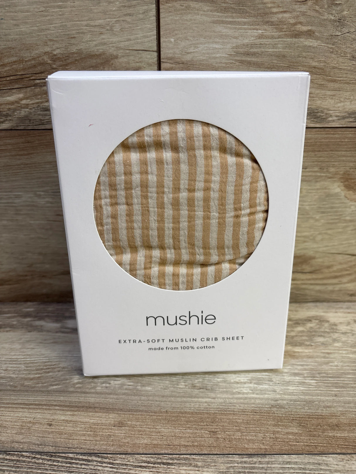 Front view of Mushie mini crib sheet showcasing natural tan stripe extra-soft muslin through round box window on wooden surface