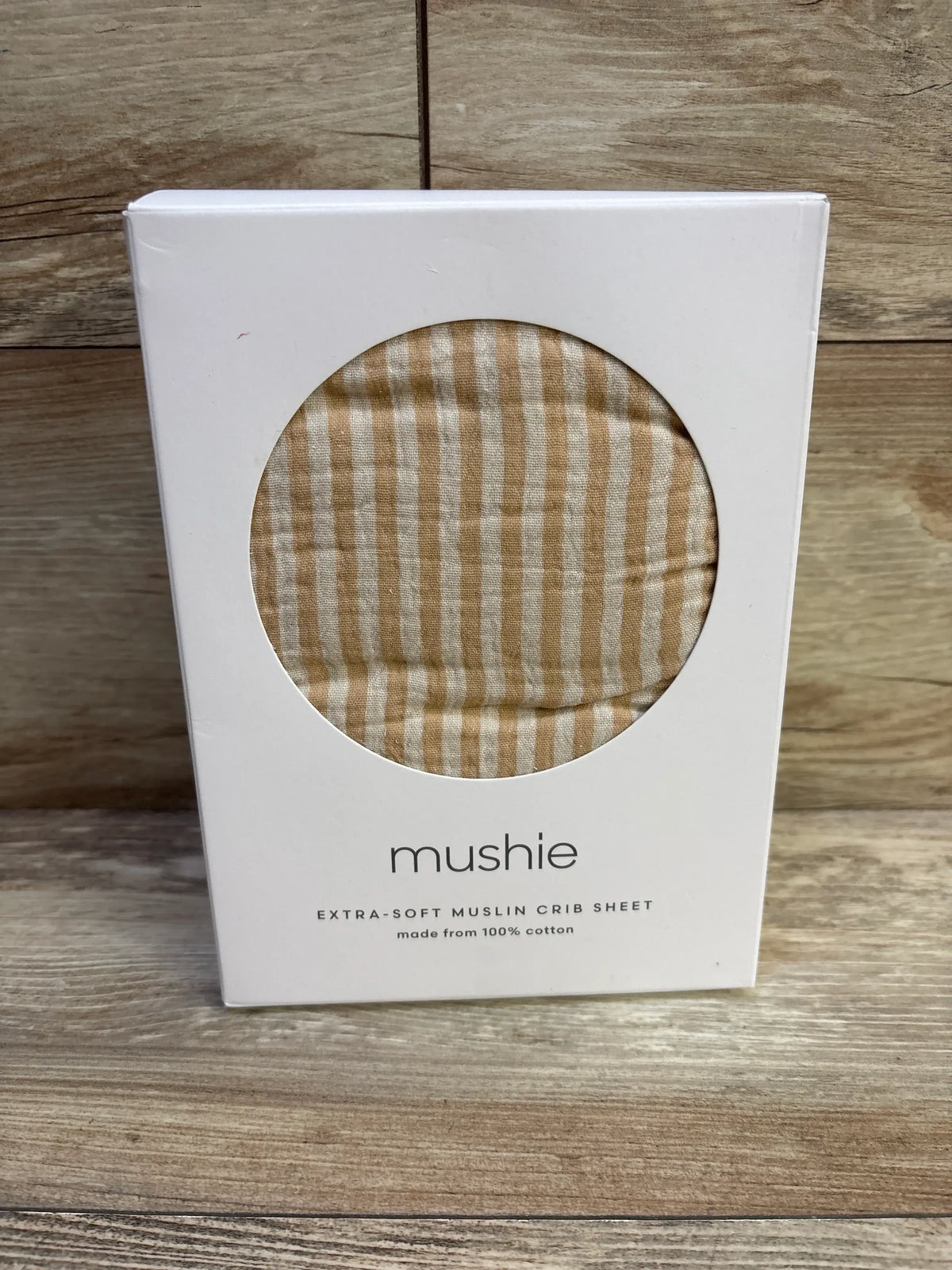 Front view of Mushie mini crib sheet showcasing natural tan stripe extra-soft muslin through round box window on wooden surface