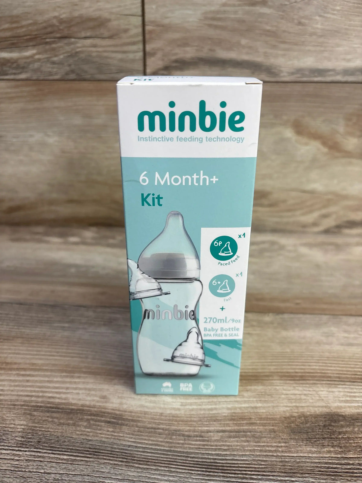 Front view of Minbie 6 Month plus feeding kit box showing bottle illustration, 270 ml size and teal and white packaging on wooden tabletop