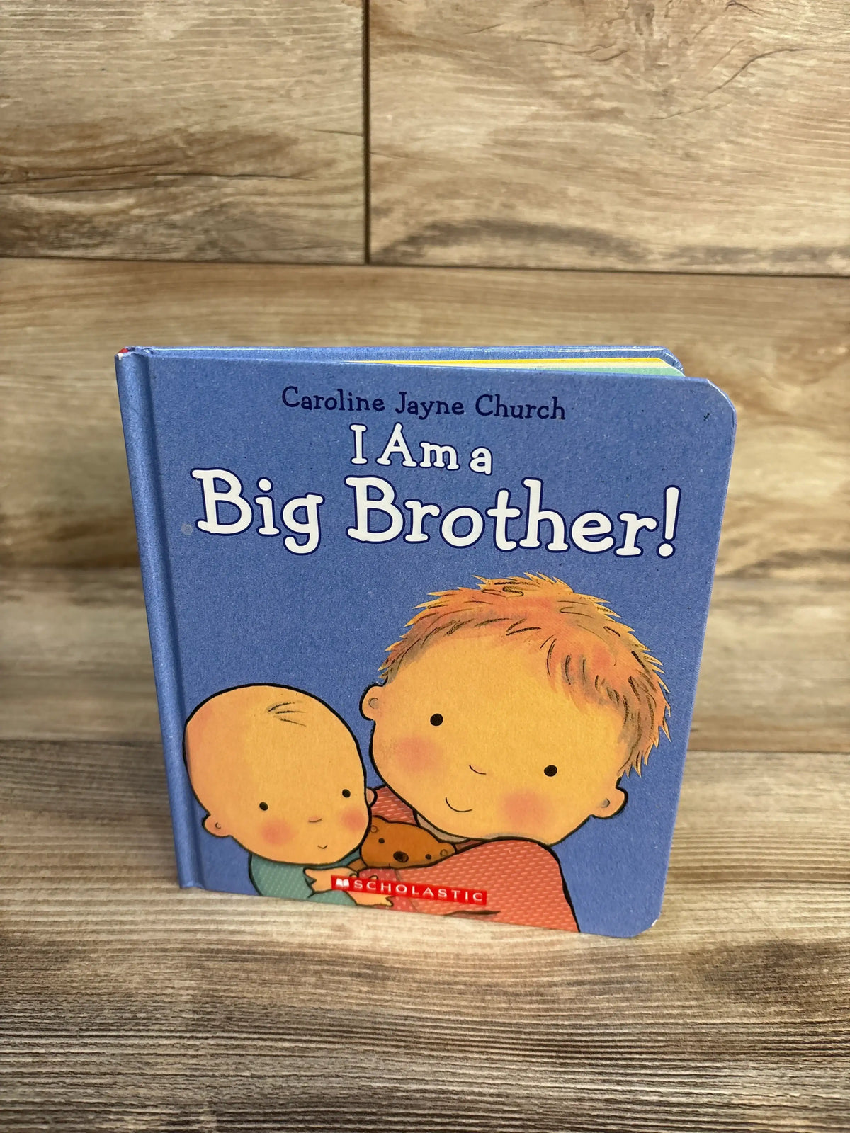 Front angled view of I Am a Big Brother Hardcover Book showcasing a blue cover with an older child hugging a baby on a wood surface