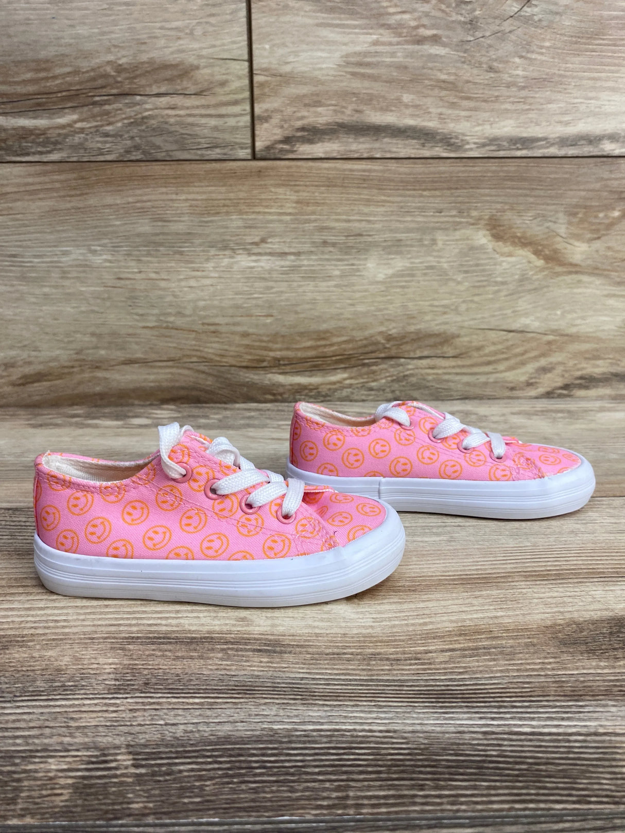 Pink kids sneakers with white laces and orange smiley face pattern on wooden floor