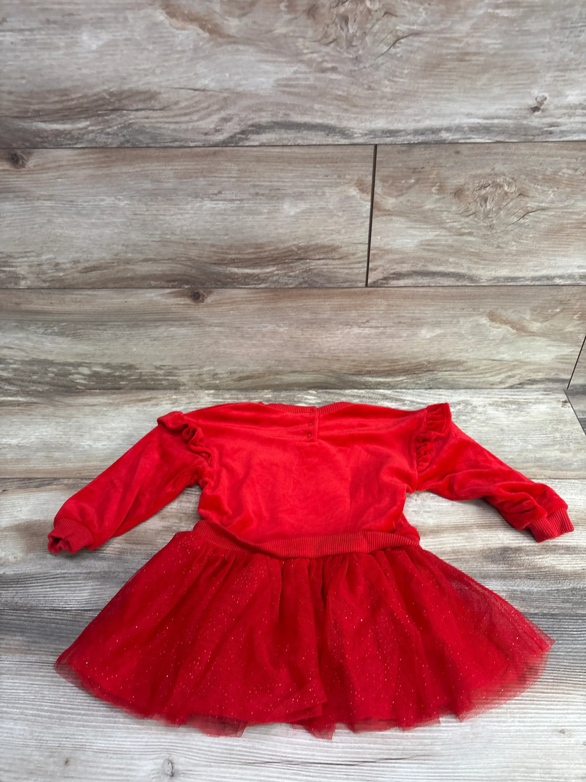 Front view of Disney Baby Minnie Mouse red velour sweatshirt dress with ruffled long sleeves and glittery tulle skirt on a wood backdrop