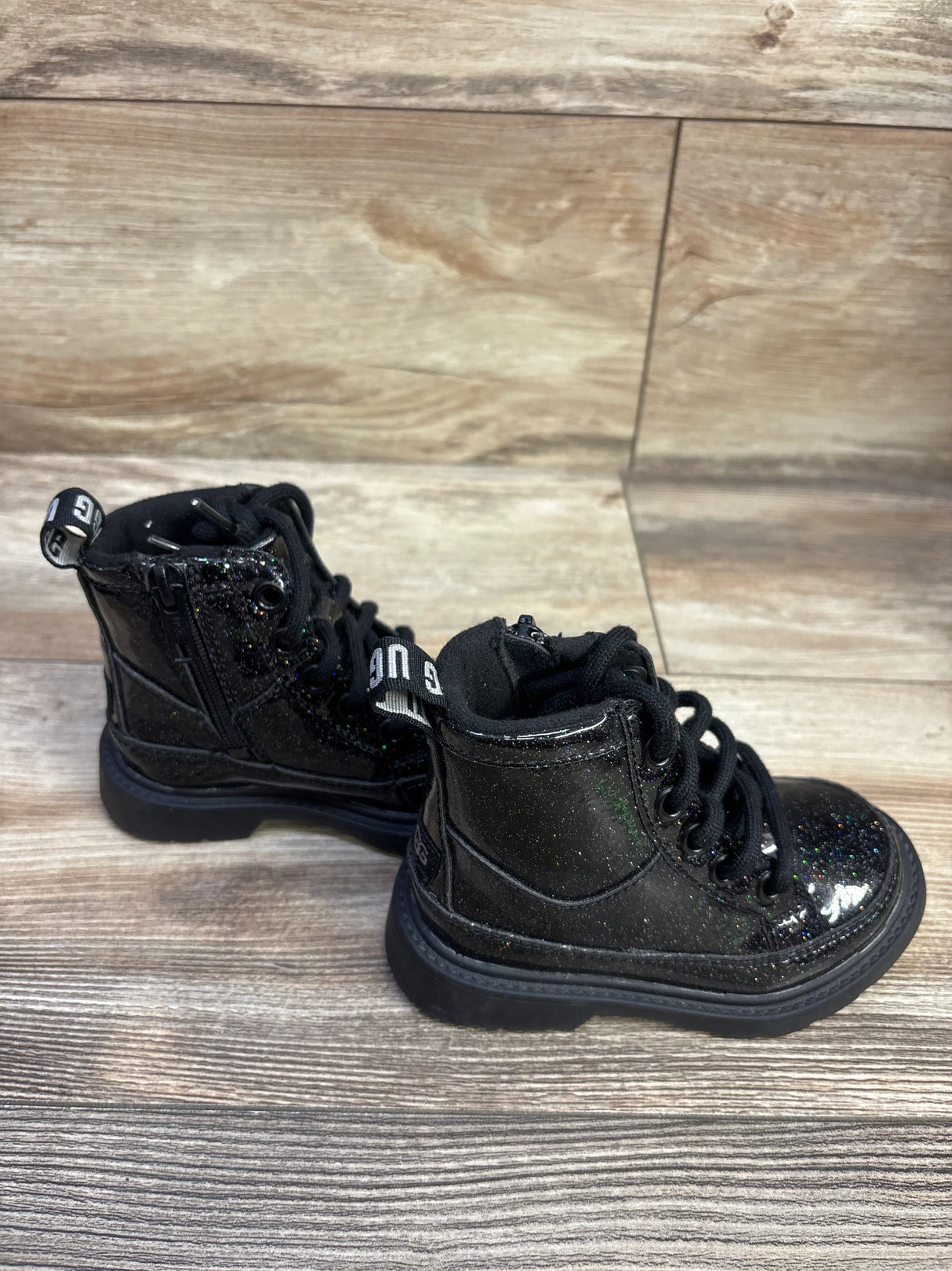 Three-quarter view of UGG Kids Robley boots with black glitter, laces, side zipper and heel tab on wood floor
