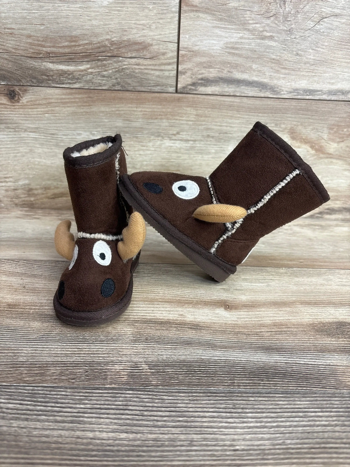 Front and side view of Lazy One kids moose slipper boots with brown faux-suede, plush lining and tan antlers on wood