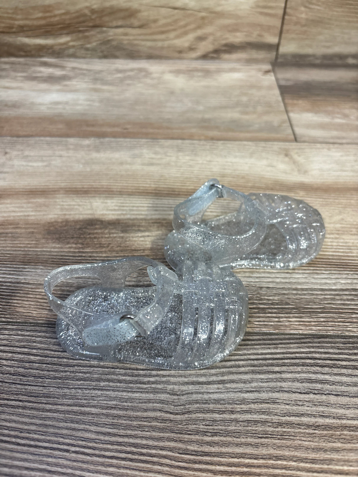Three-quarter view of Old Navy clear glitter jelly toddler sandals showing caged toe and ankle strap on wood background