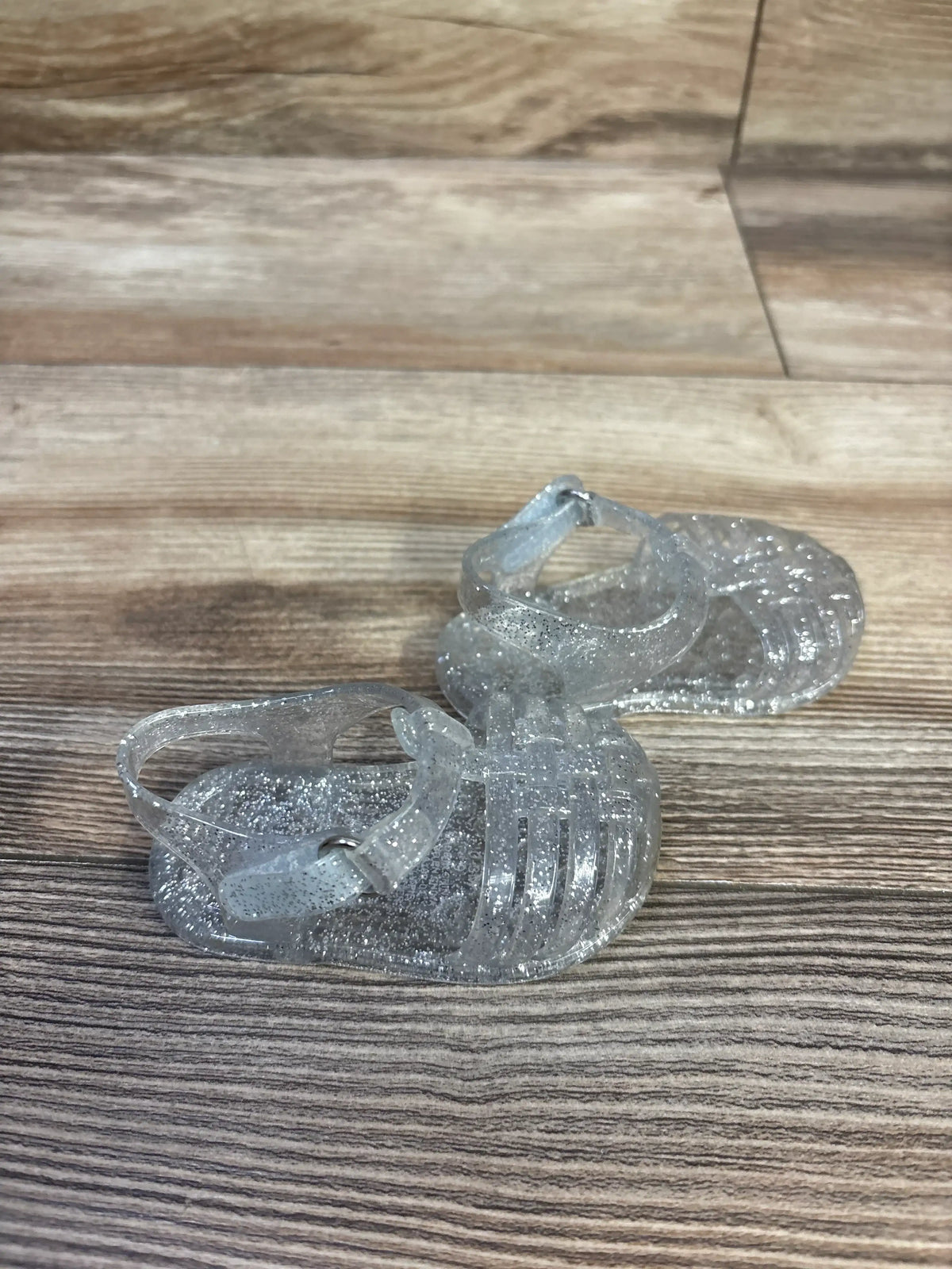 Three-quarter view of Old Navy clear glitter jelly toddler sandals showing caged toe and ankle strap on wood background