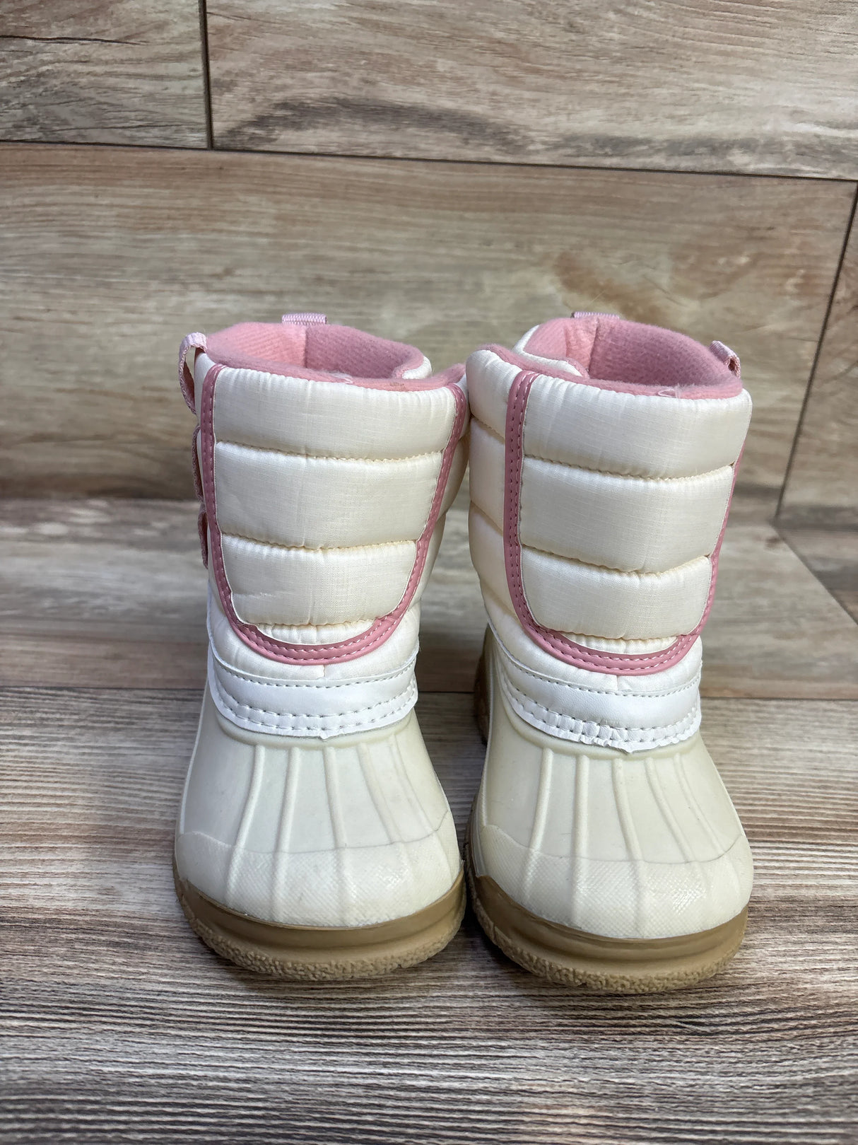 Front view of Oshkosh toddler winter boots showing ivory quilted upper, pink trim and rubber toe and sole on wood background