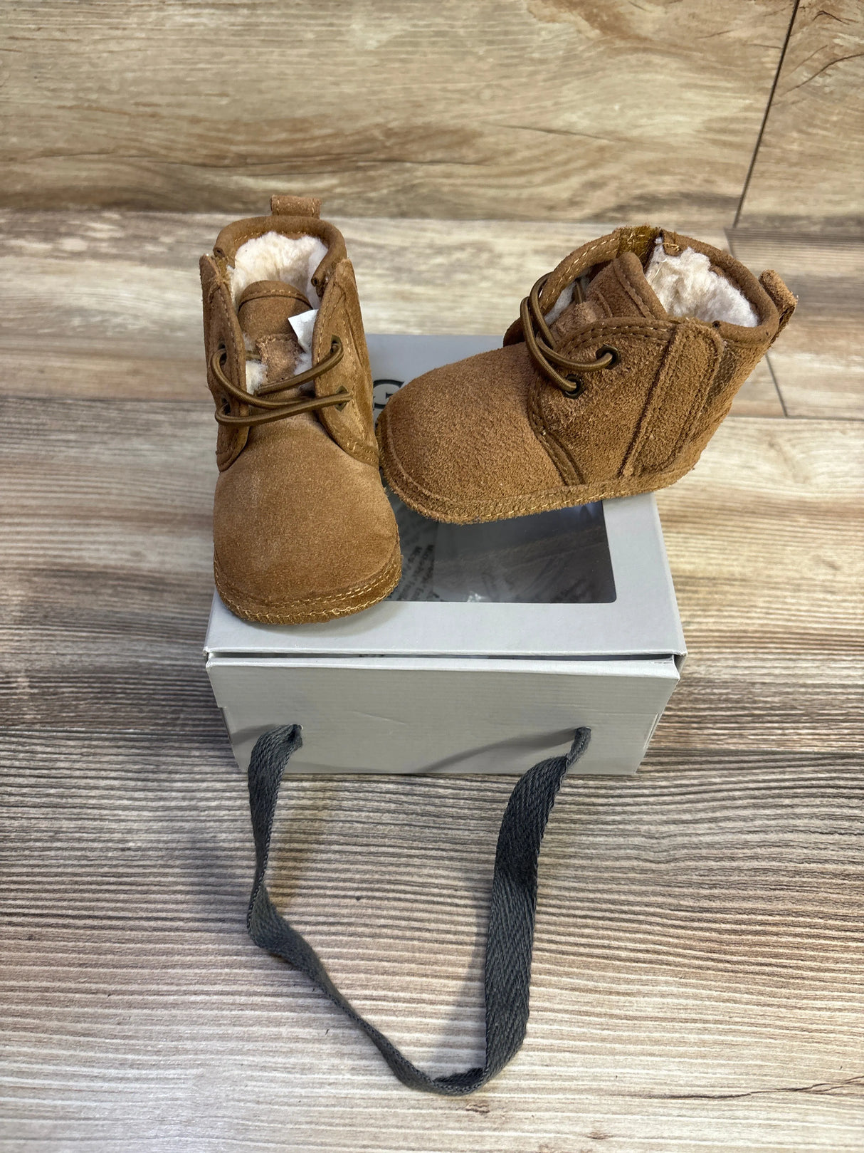Front and side view of UGG Baby Neumel chestnut booties with shearling lining and laces on a box on wood background