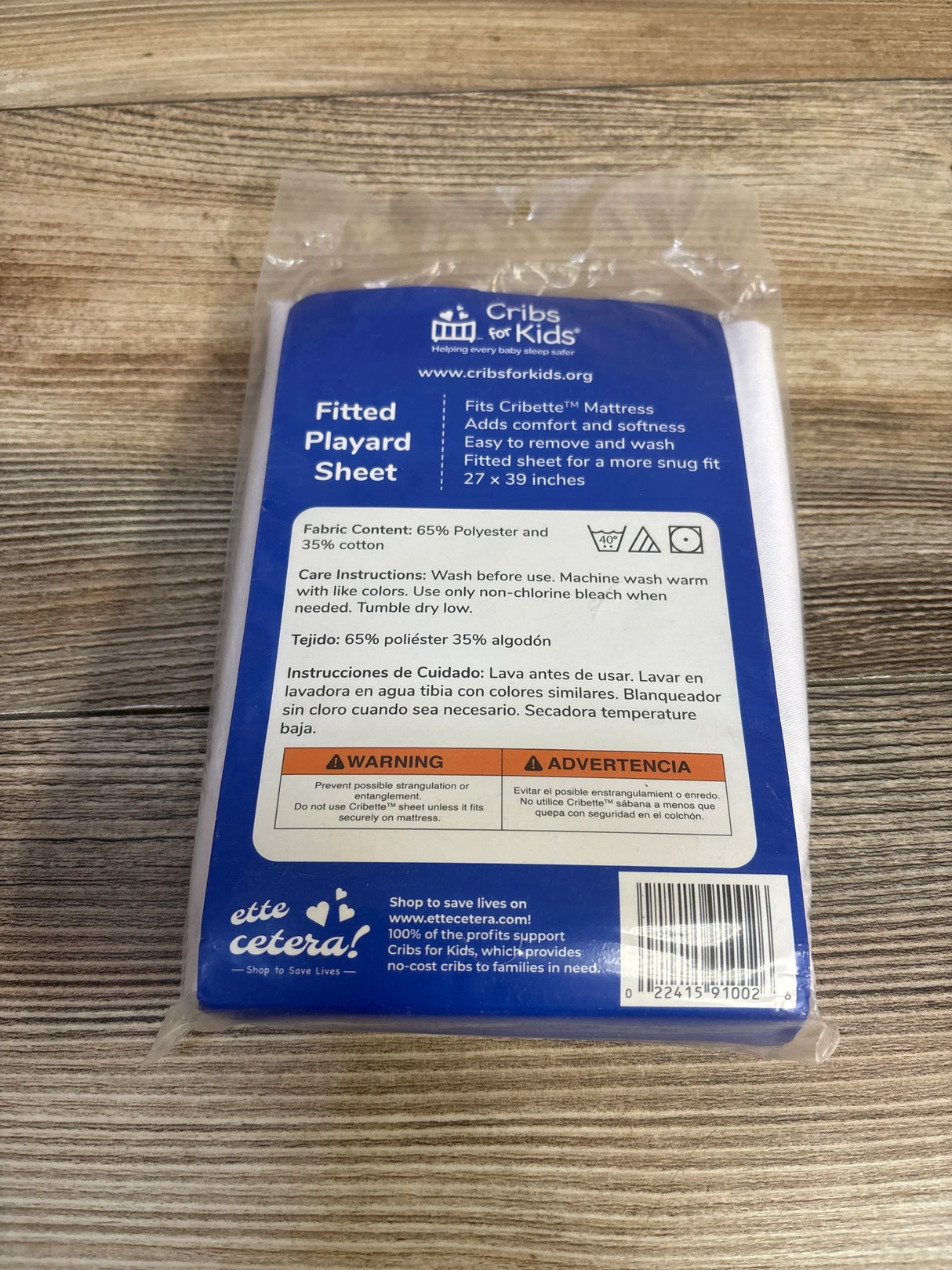 Top-down view of Cribs for Kids fitted playard sheet in sealed packaging showing blue label and size 27 x 39 in on wood table