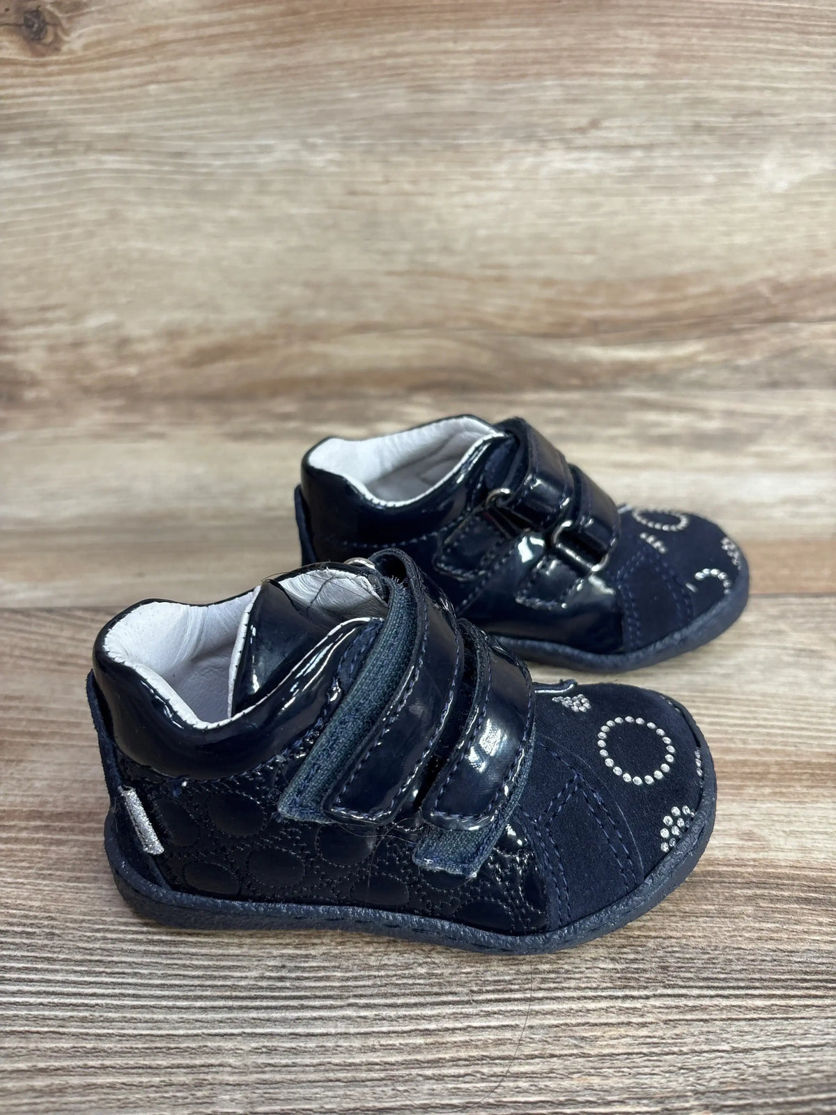 Three-quarter view of navy Melania baby sneakers with double velcro straps and circular rhinestone accents on wooden surface