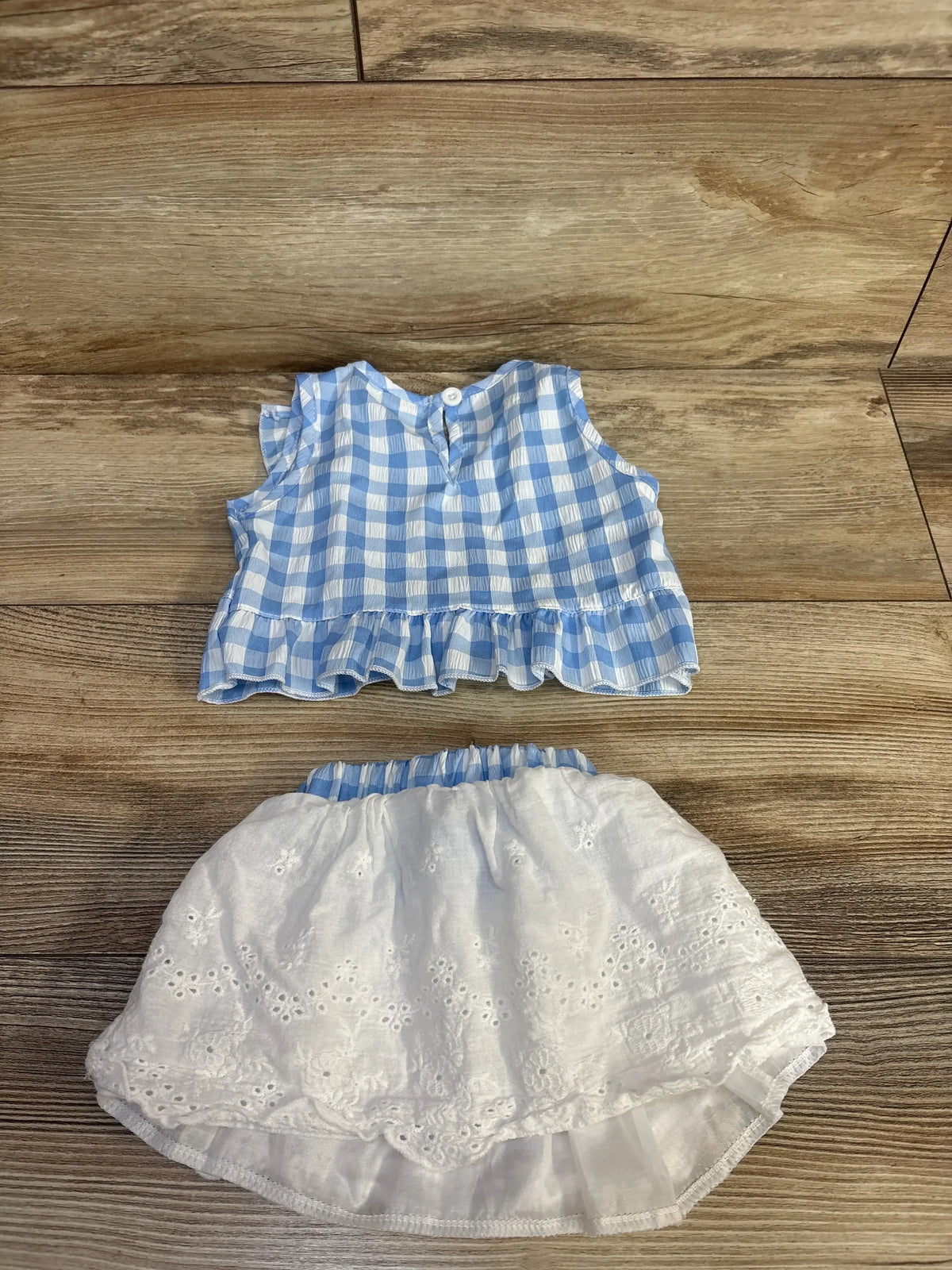 Flat lay of 2-piece baby outfit showcasing light blue gingham ruffle blouse and white eyelet skirt with wood background